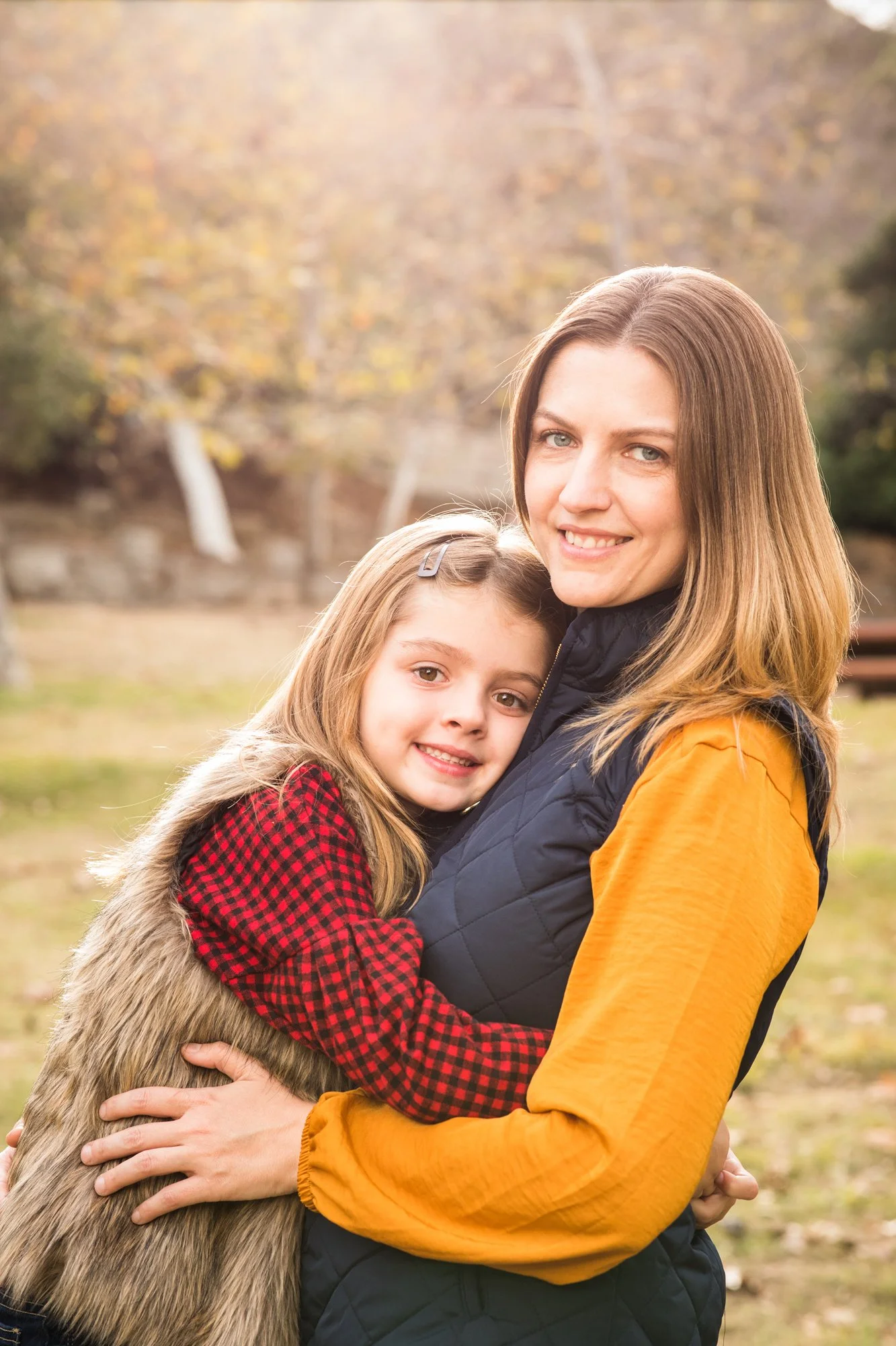 A woman and a girl hugging outdoors during autumn, with trees and fallen leaves in the background.