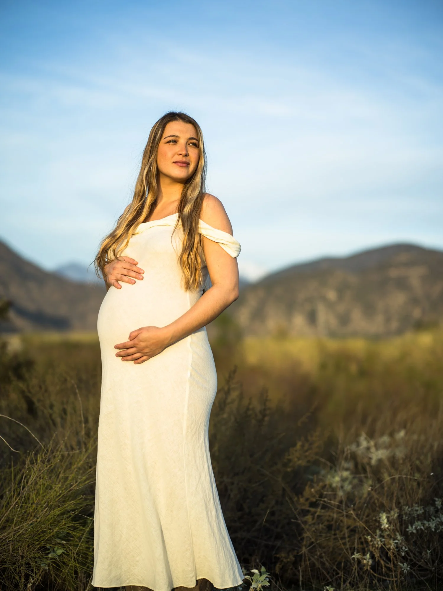 Pregnant woman standing outdoors in a field, wearing a cream-colored dress, with mountains in the background and a blue sky.
