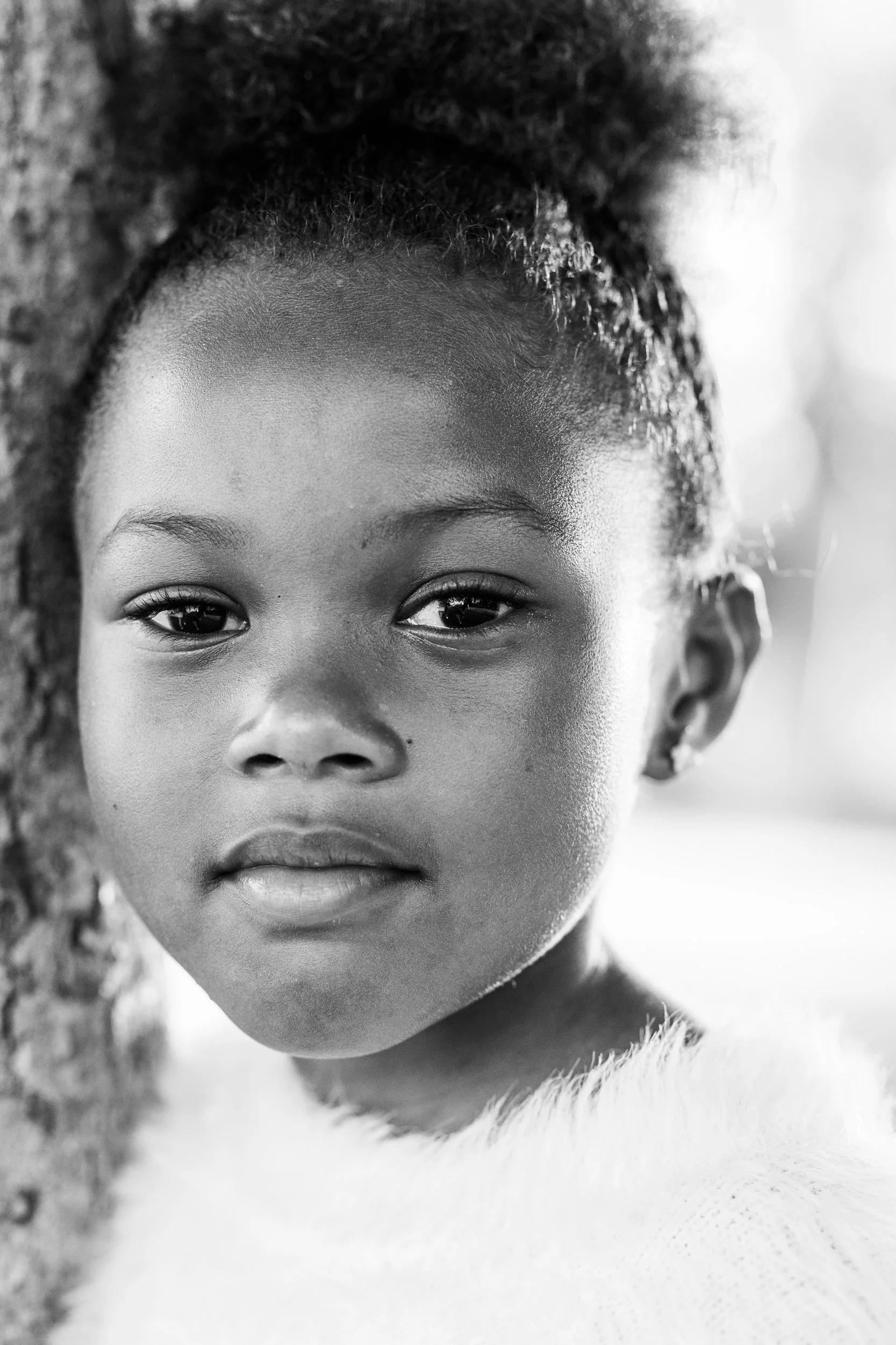 Close-up black and white portrait of a young girl with natural hair, leaning against a tree.