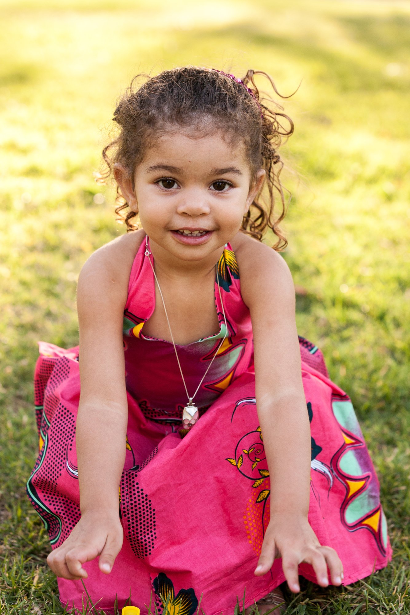 A young girl with curly hair, wearing a colorful pink dress with yellow, black, and green patterns, is sitting on grass outdoors, smiling at the camera.