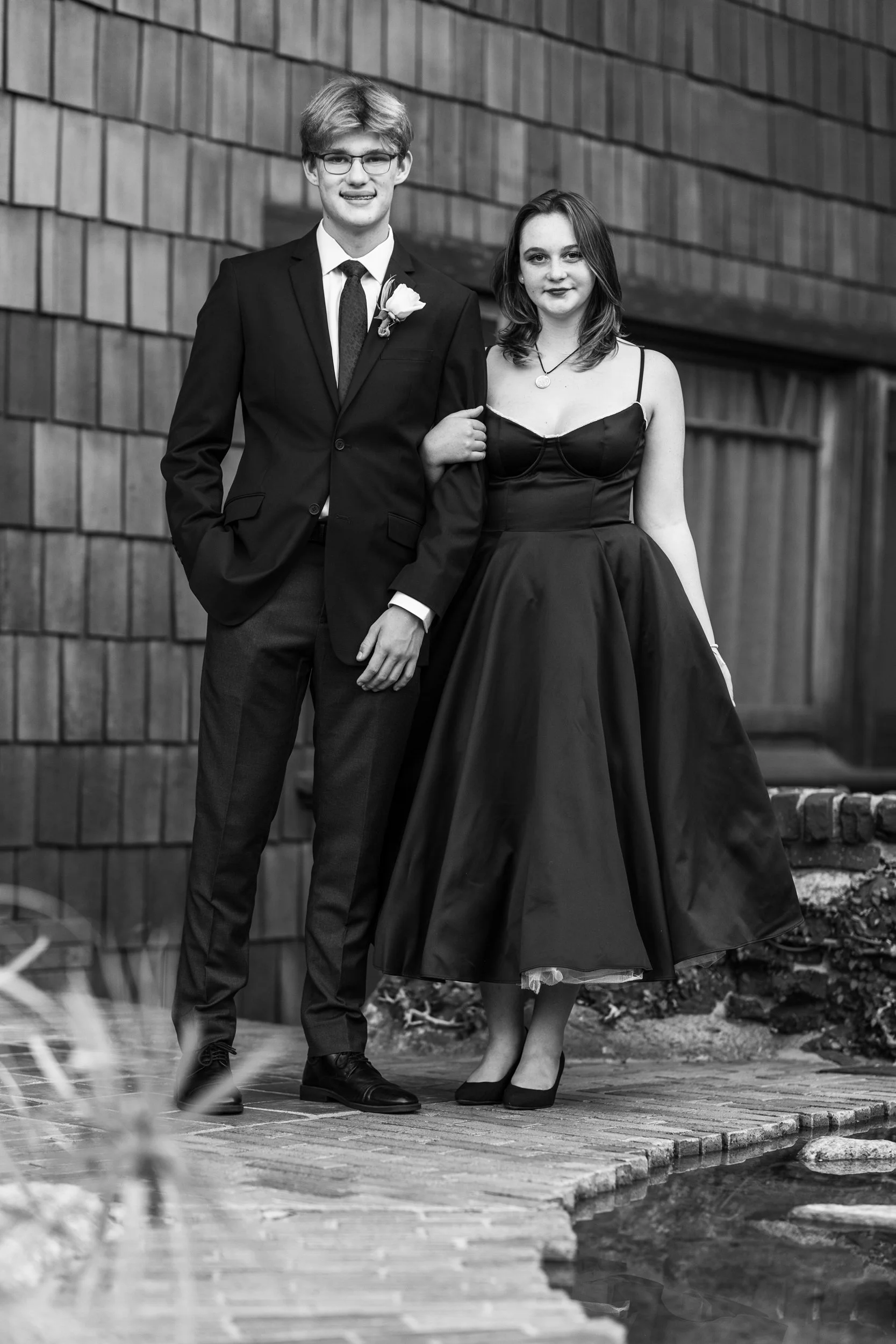 A young man and woman dressed in formal attire, standing arm in arm outdoors in front of a building with shingle siding.