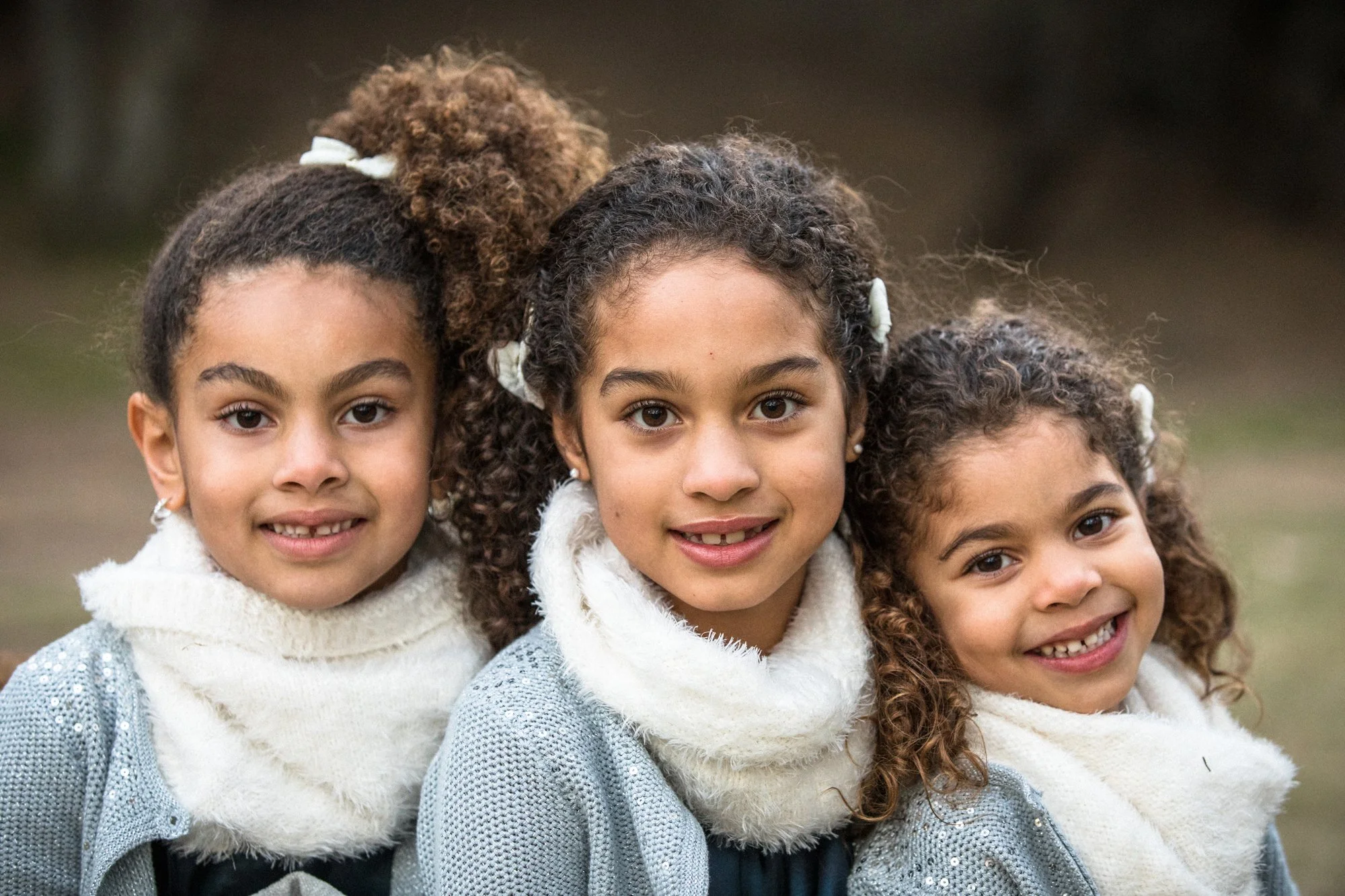 Three young girls with curly hair, wearing sweaters and scarves, smiling outdoors.
