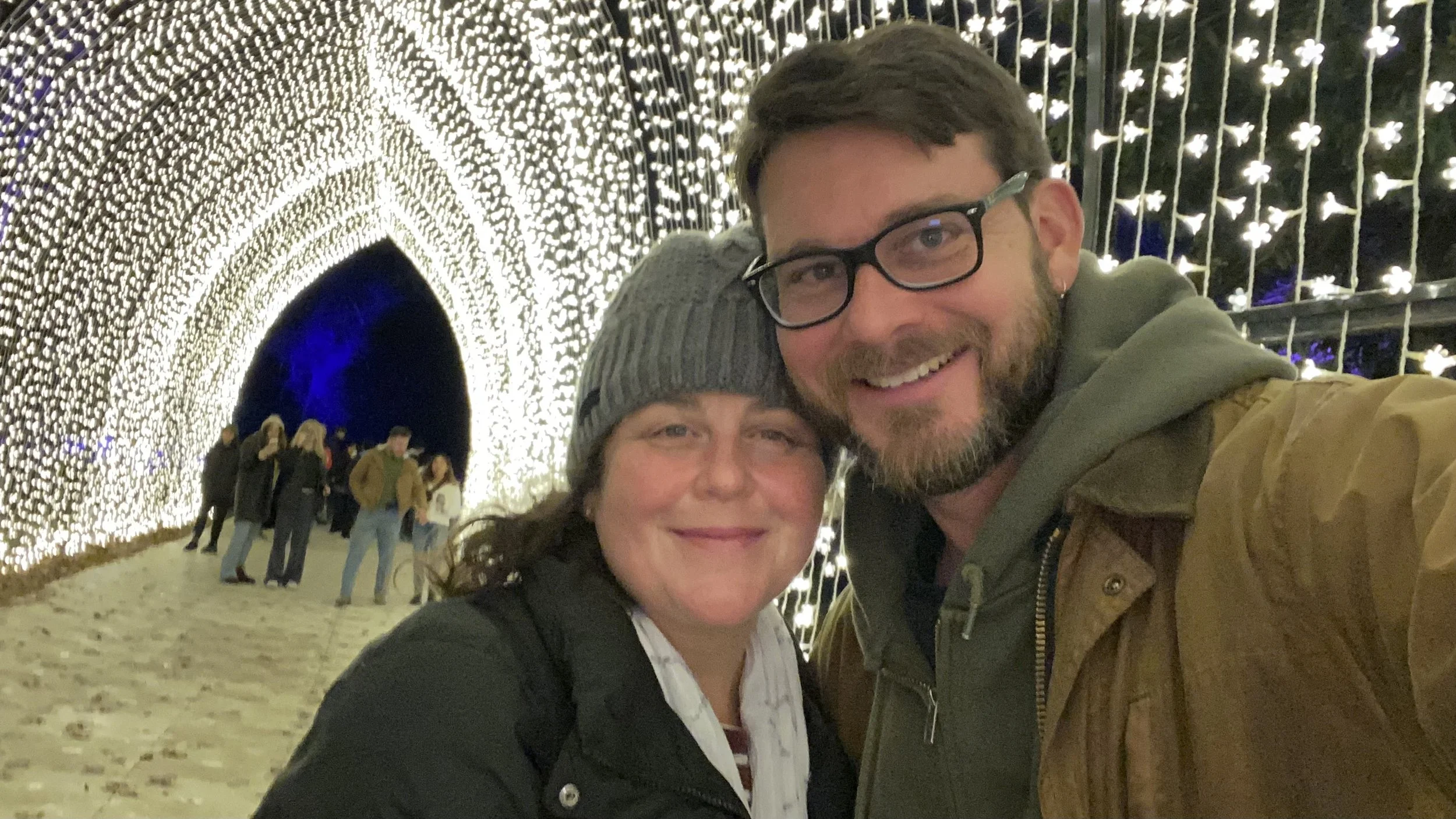 A smiling man and woman taking a selfie together in front of an illuminated tunnel with many small white lights, with other people in the background.
