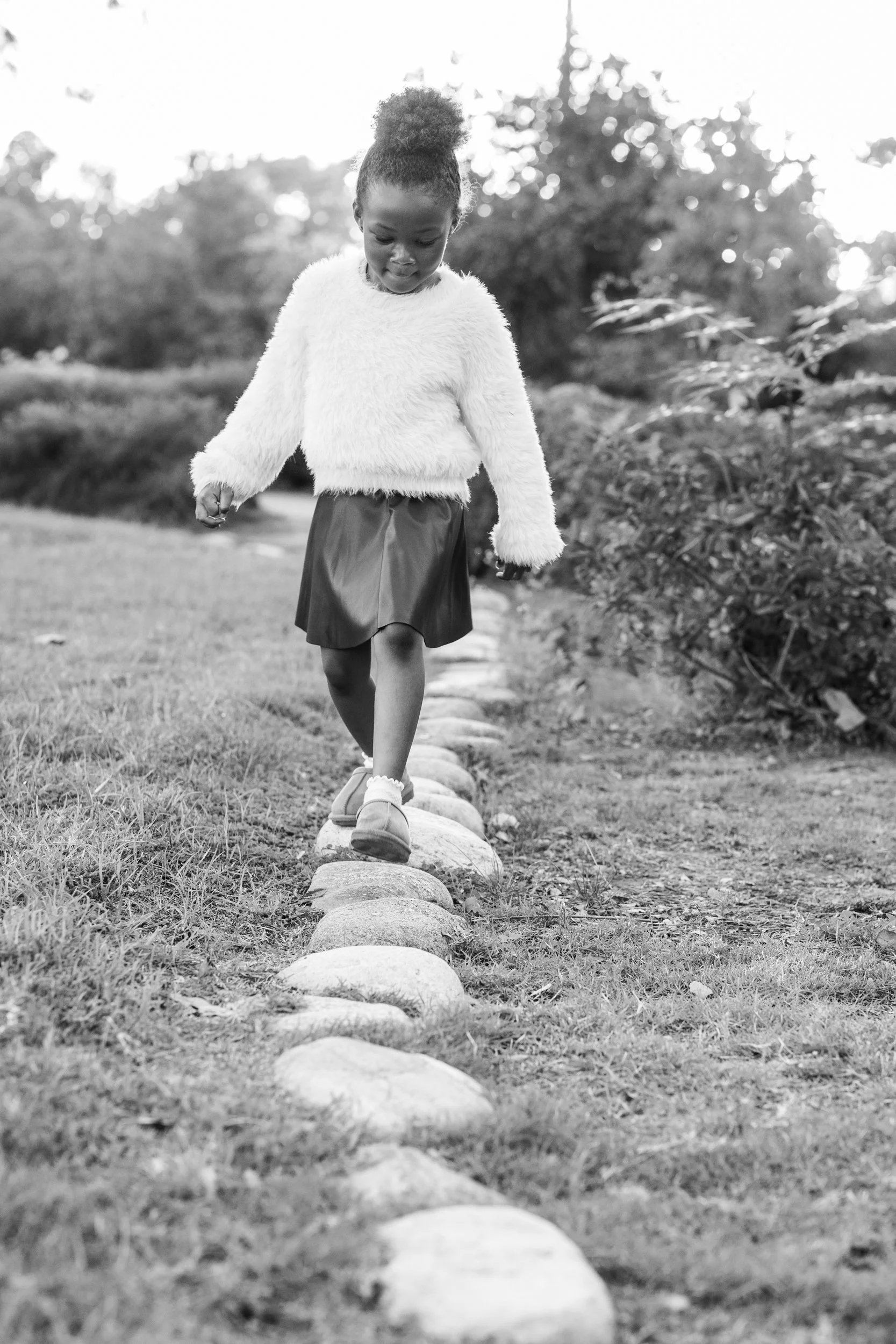 A young girl walking on a line of stones in a park, wearing a fluffy sweater, a skirt, and shoes.