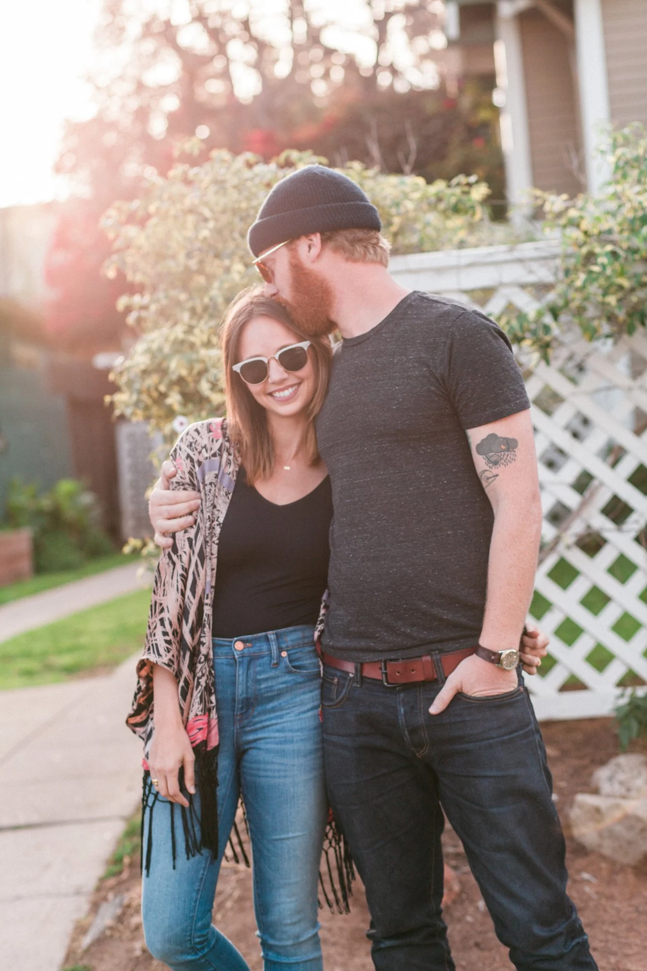 A young man and woman standing outdoors on a sidewalk, smiling and embracing each other. The woman is wearing sunglasses, a black top, blue jeans, and a patterned shawl. The man is wearing a beanie, black t-shirt, jeans, and has arm tattoos. The back