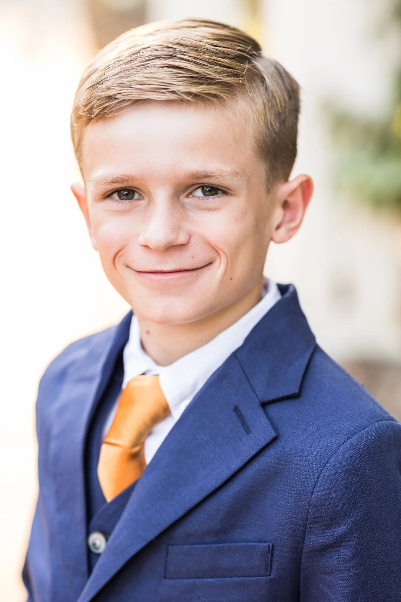 Young boy in a blue suit jacket, white shirt, and orange tie smiling outdoors.