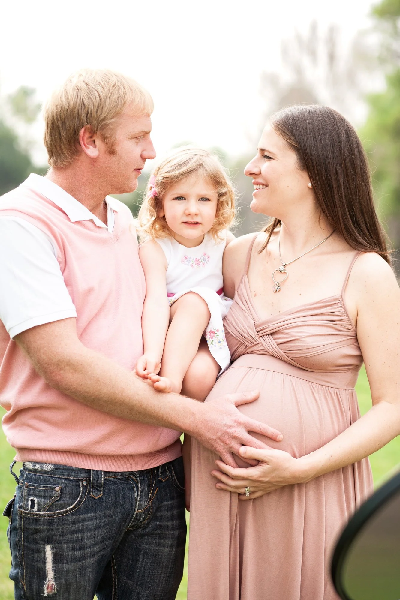 A family outdoors with a pregnant woman, her partner, and their young daughter. The woman is gently holding her pregnant belly, while the man and daughter look at her. The scene appears warm and joyful.
