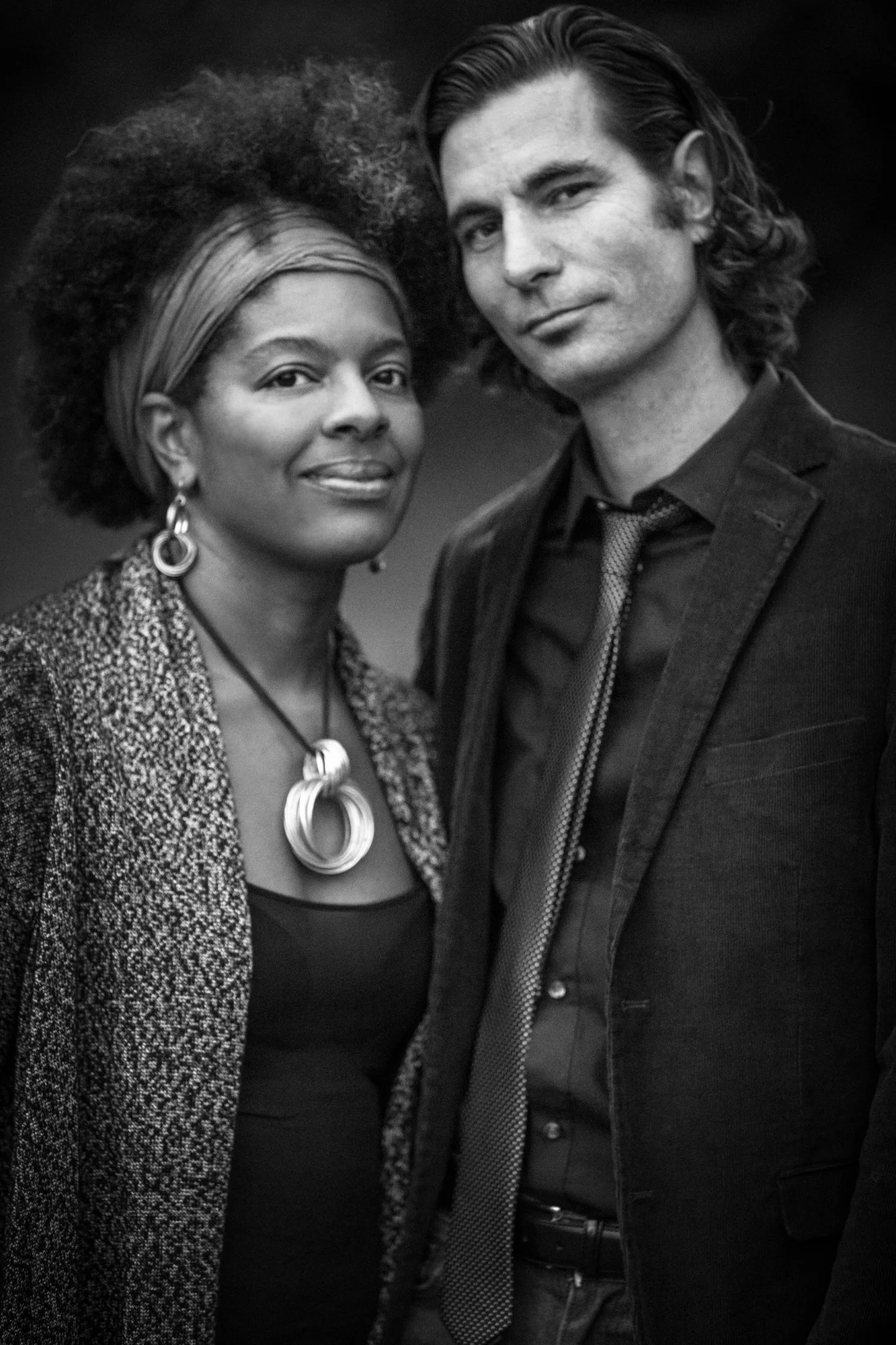 Black and white photo of a woman with curly hair and a man with wavy hair, both dressed in formal attire, standing close together and looking at the camera.