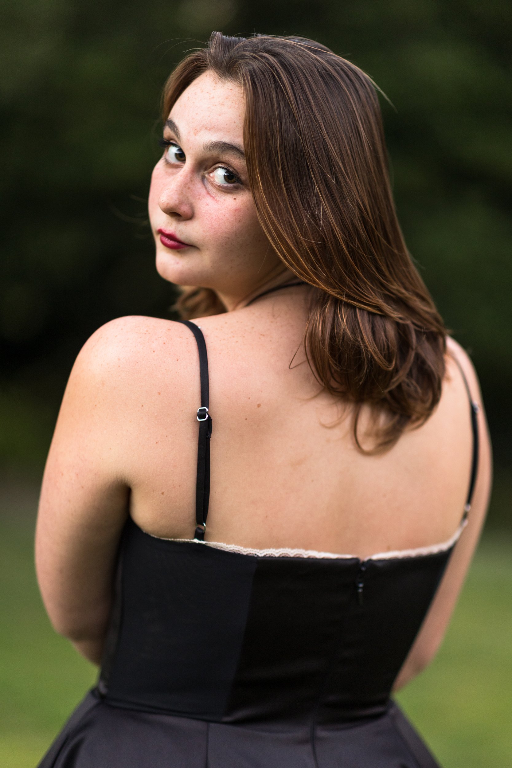 A young woman with shoulder-length brown hair and light skin, wearing a black spaghetti strap dress, standing outdoors with green foliage in the background, looking over her shoulder.
