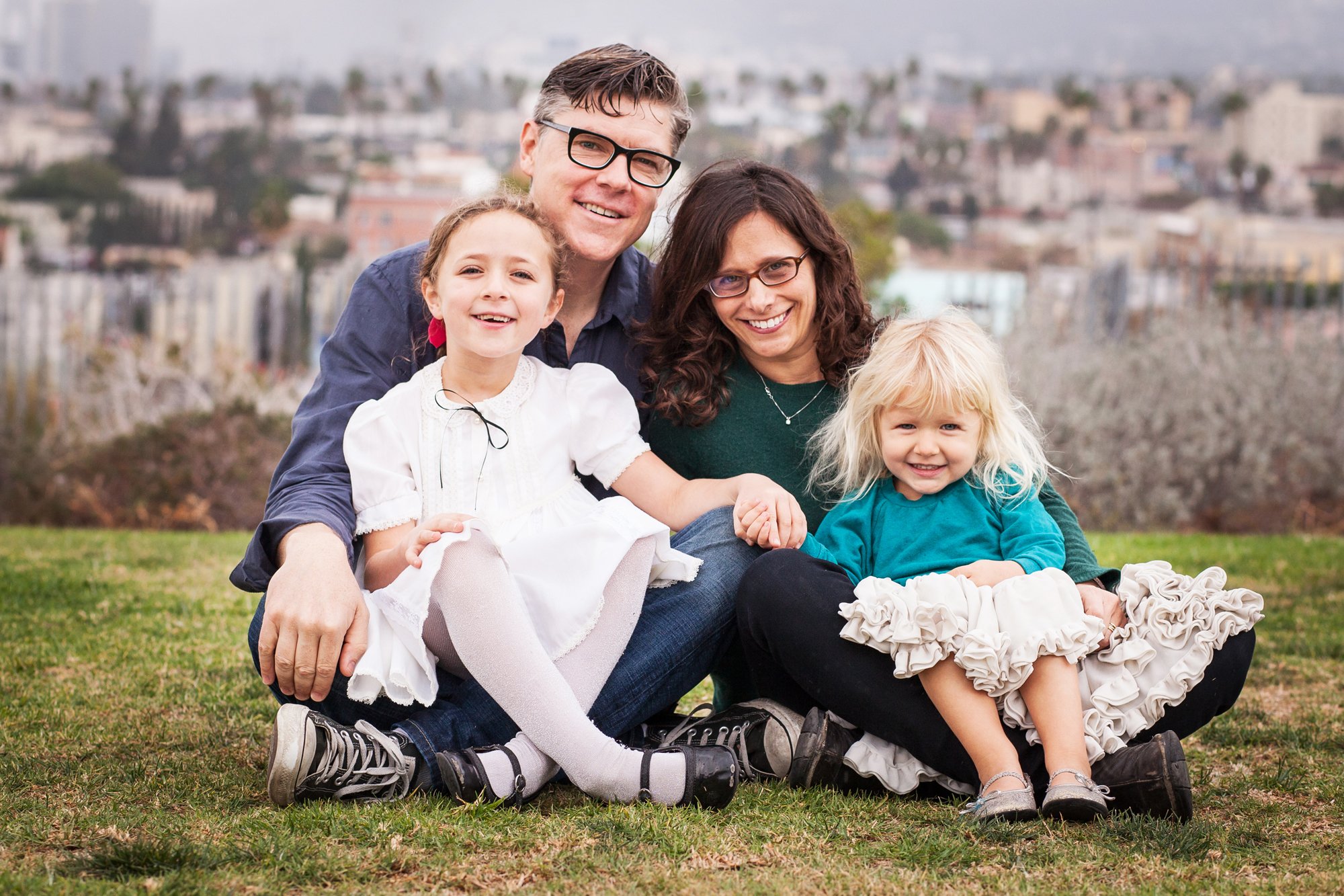 A happy family of four sitting on grass with cityscape in the background, including a man, woman, and two young girls.