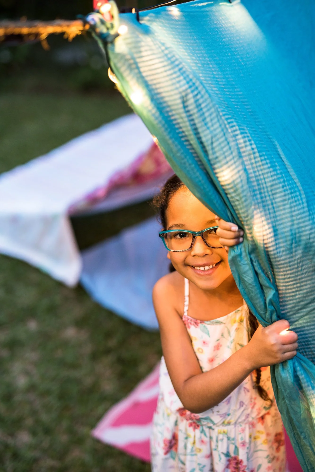A young girl with glasses smiling and peeking out from behind a blue curtain at an outdoor event during dusk.