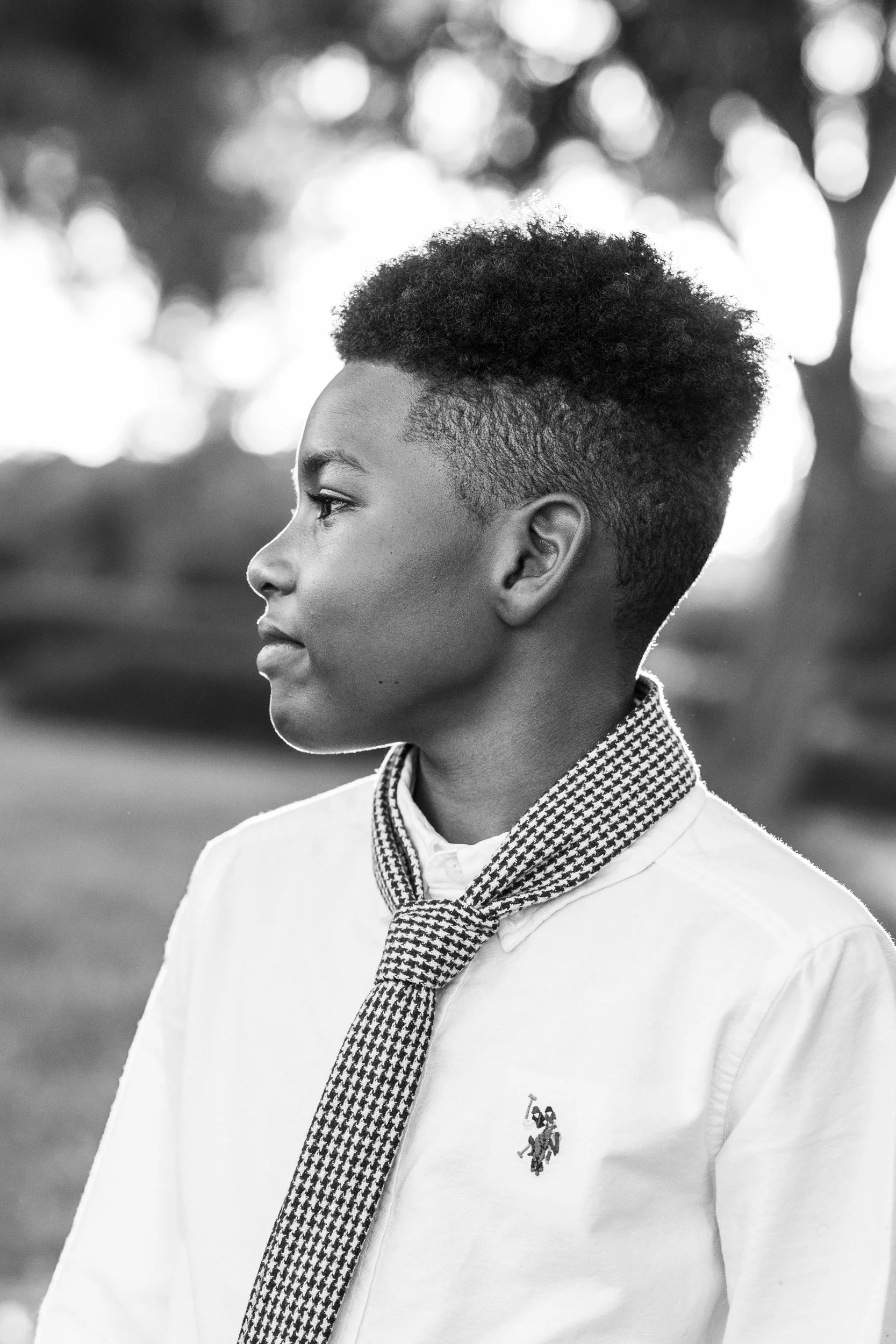 Black and white profile photo of young man with short curly hair, wearing a collared shirt and a patterned scarf, outdoors with blurred trees in the background.