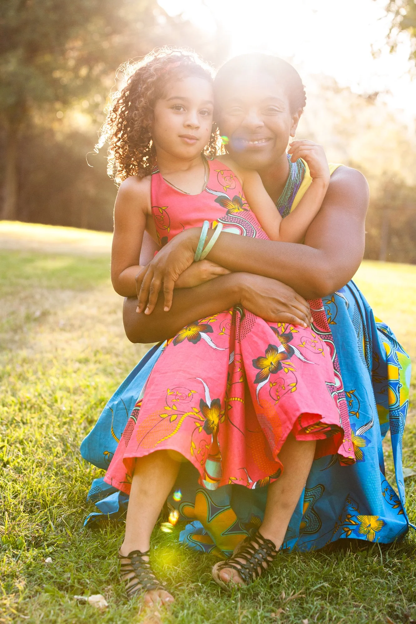 A woman and a young girl are outside in a grassy area during sunset, embracing and smiling at the camera. The woman has short hair and is wearing a blue wrap skirt with yellow and green patterns, while the girl has curly hair and is dressed in a pink