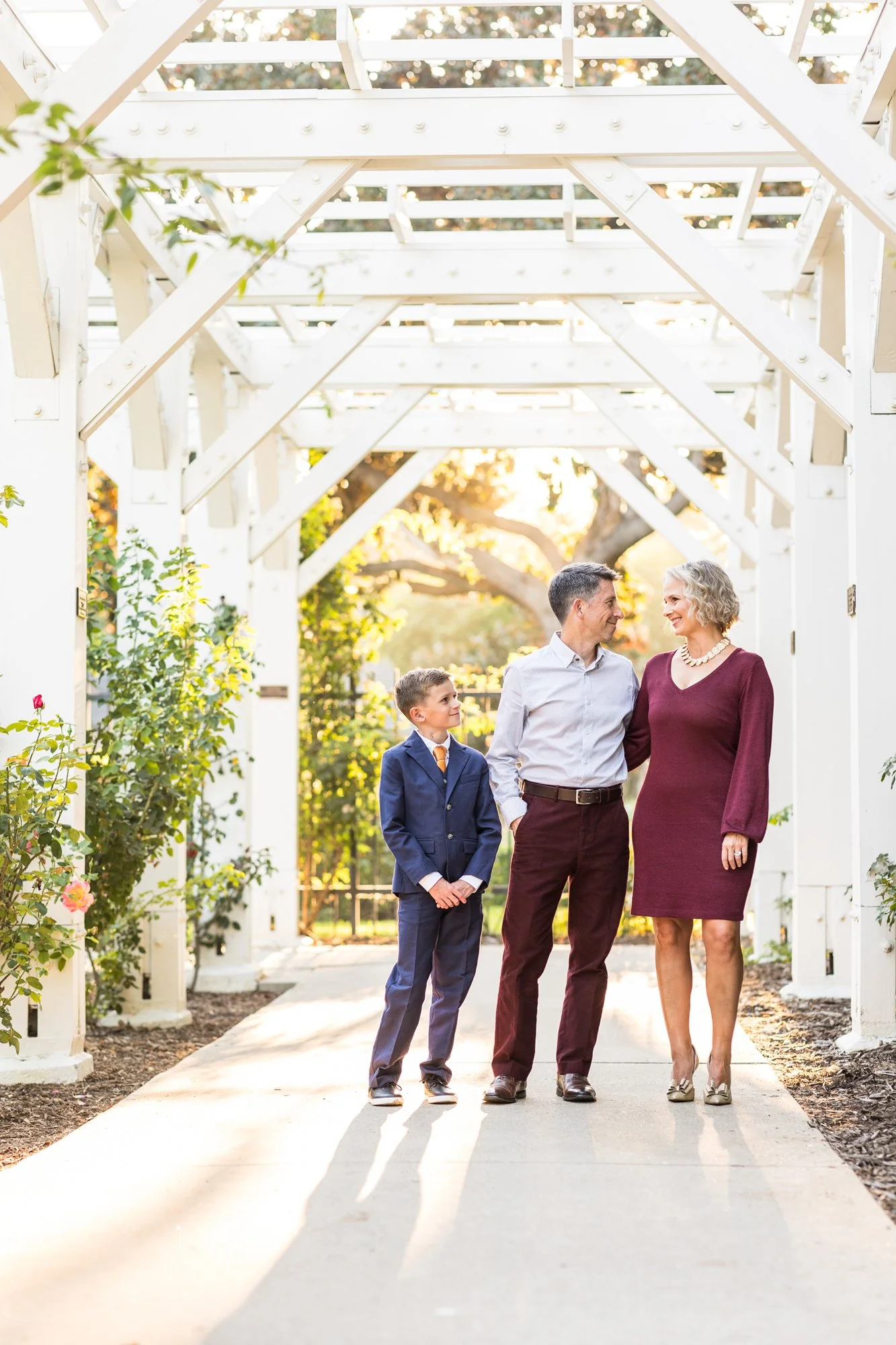 A family of three, a man, a woman, and a young boy, walking together under a white arbor in a garden during sunset, smiling and enjoying time together.