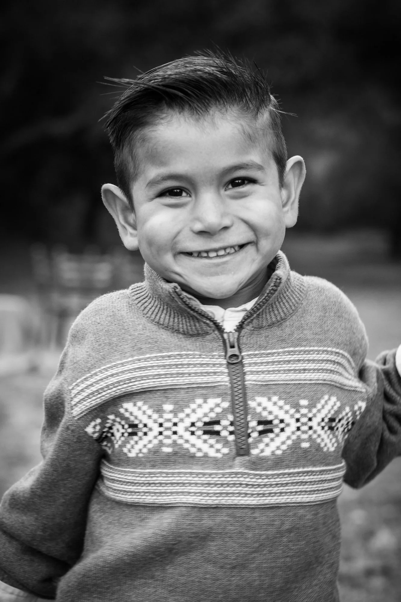 Young boy smiling outdoors, wearing a patterned sweater, in black and white.