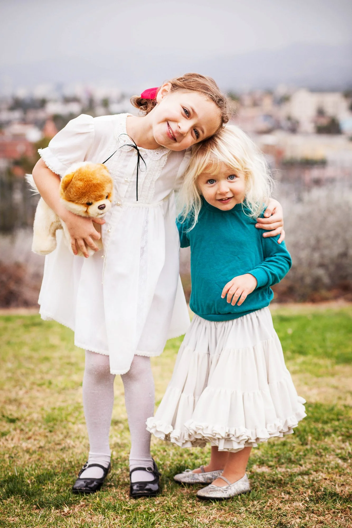 Two young girls, one with light blonde hair and the other with light brown hair, smiling outdoors with arms around each other. The girl with blonde hair is wearing a teal top and a cream-colored tiered skirt, while the girl with brown hair is holding