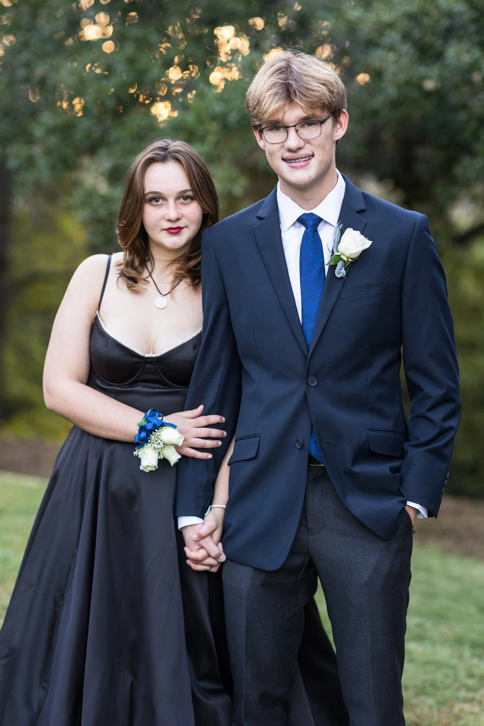 A young couple dressed in formal attire standing outdoors. The woman is wearing a black dress with spaghetti straps and a corsage on her wrist, and the man is in a navy suit with a white shirt and tie, with a boutonniere on his lapel. They are holdin
