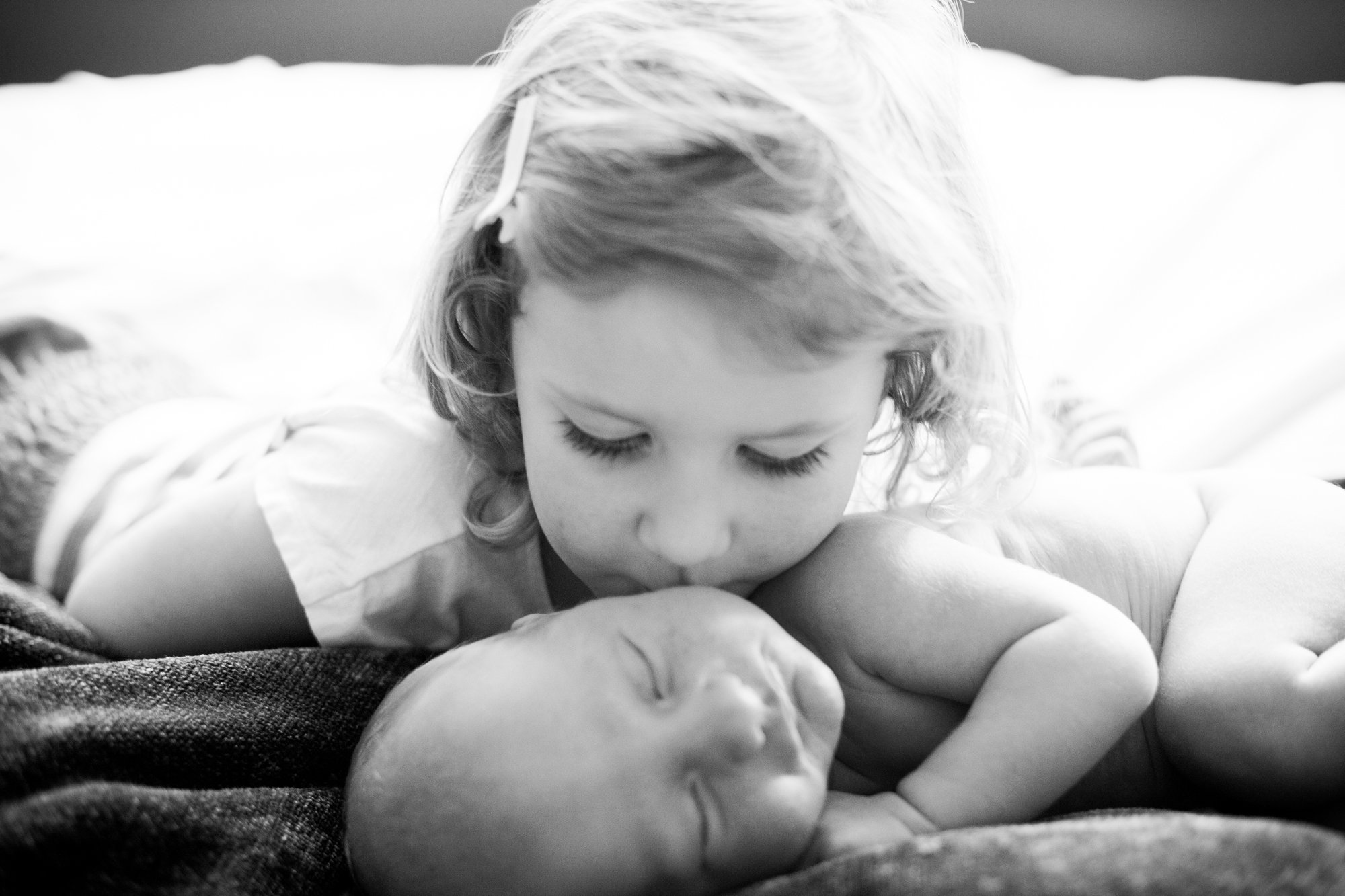 A young girl with curly hair kisses a sleeping newborn baby on the forehead.