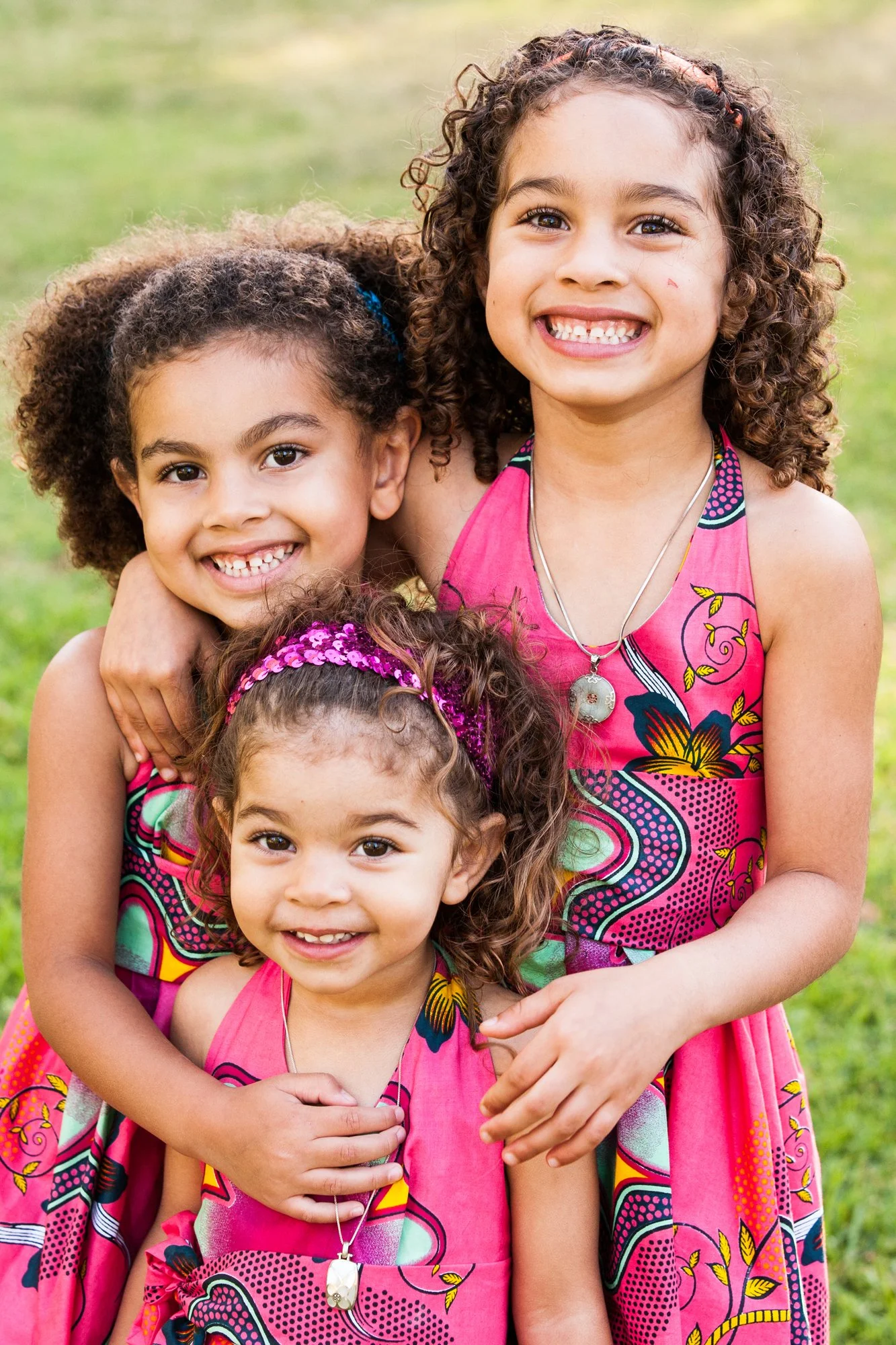 Three young girls with curly hair smiling outdoors, wearing matching pink dresses with colorful patterns and necklaces.