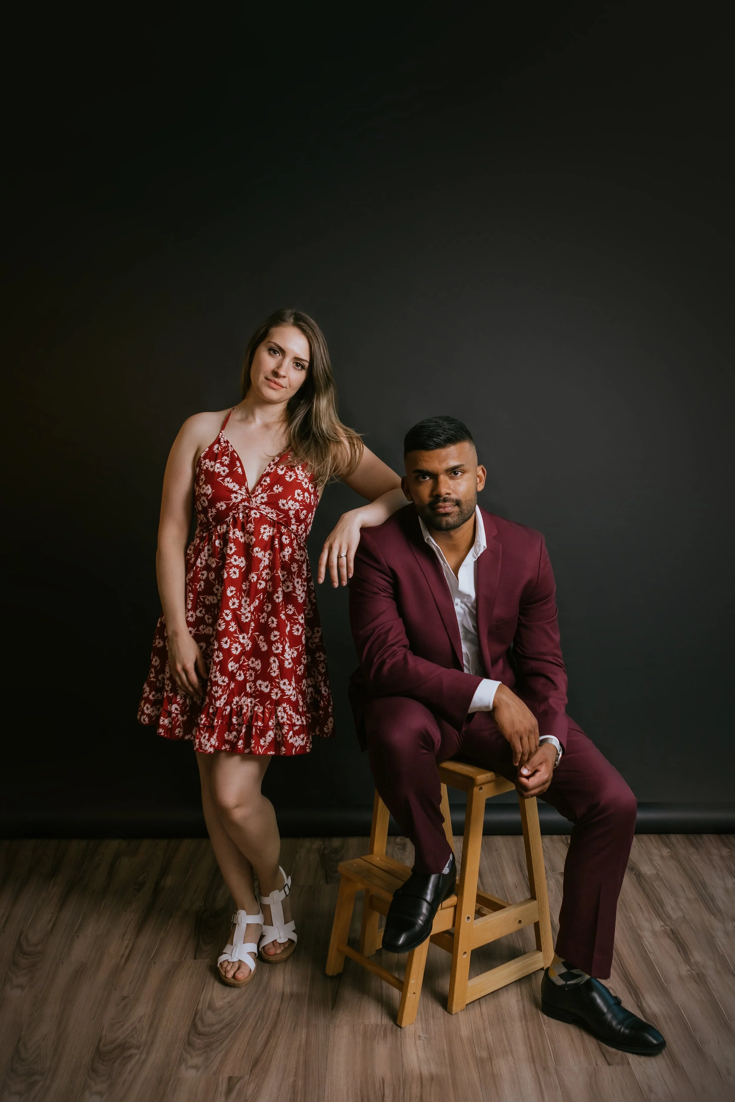 A woman in a red floral dress standing next to a man in a maroon suit sitting on a wooden chair against a black background.