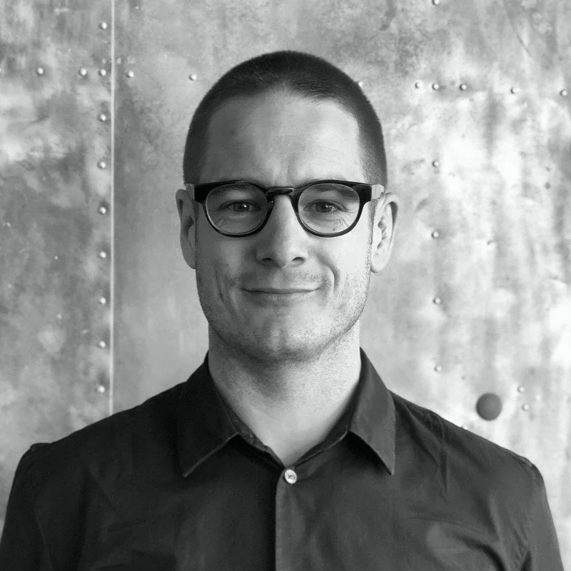 Black and white portrait of a man with short hair, wearing glasses and a collared shirt, smiling slightly, standing against a metallic textured background.