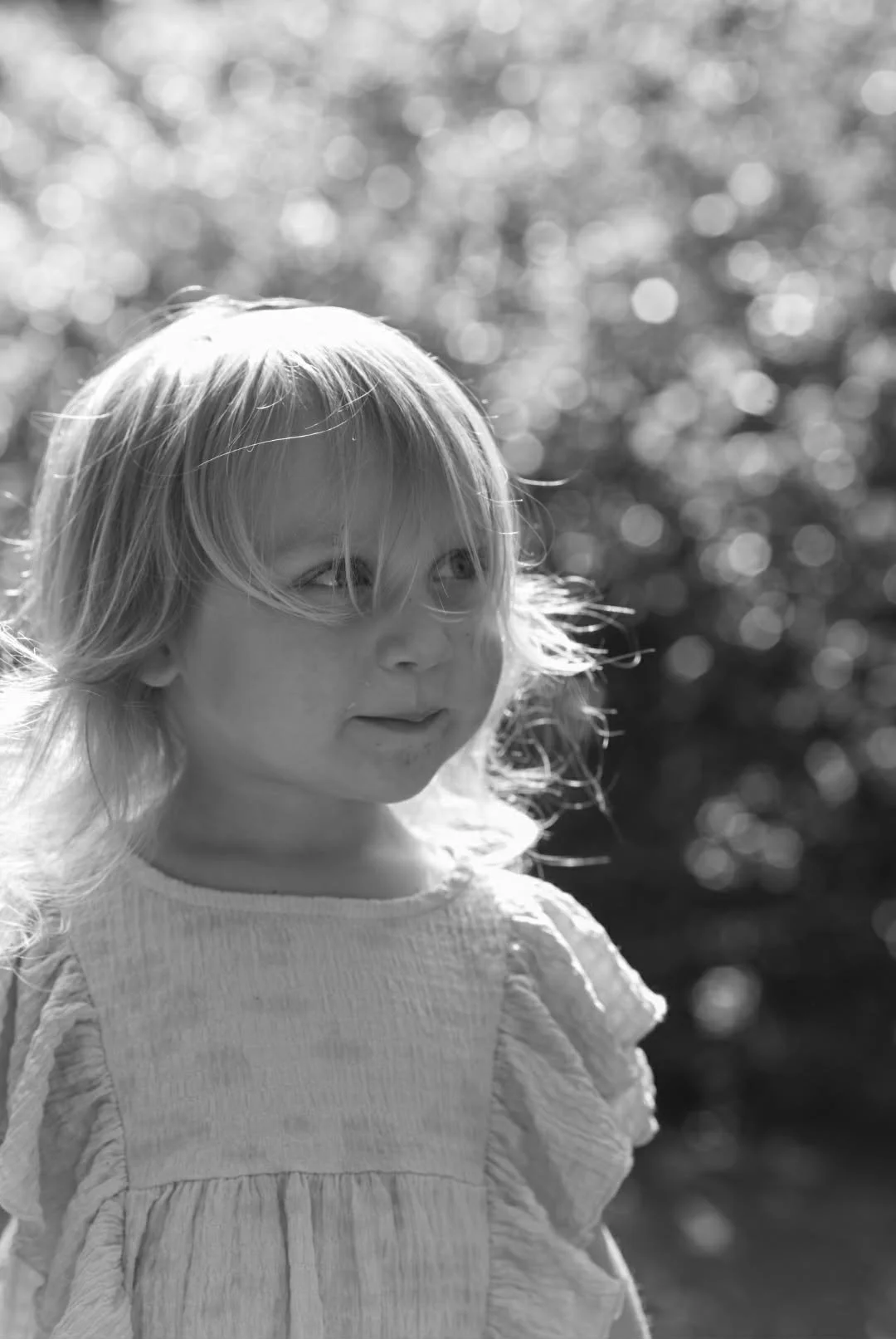 A black and white photo of a young girl with long, tousled hair, wearing a textured, light-colored top. She is outdoors with sunlight and blurred bokeh background.