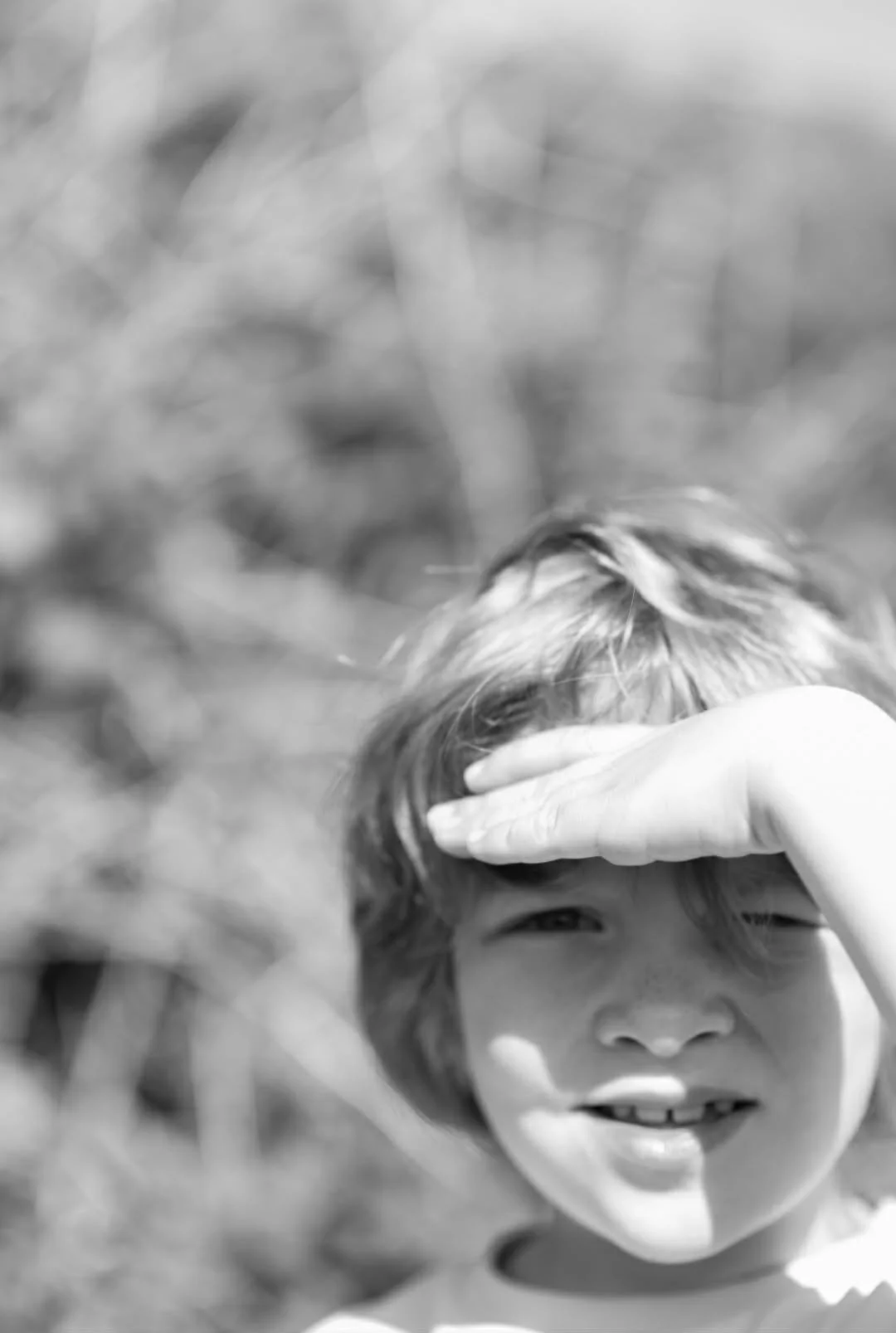 A black and white photo of a young child with short hair, holding their hand up to their forehead as if shielding eyes. The child is smiling slightly and looking toward the camera, with a blurred natural outdoor background.