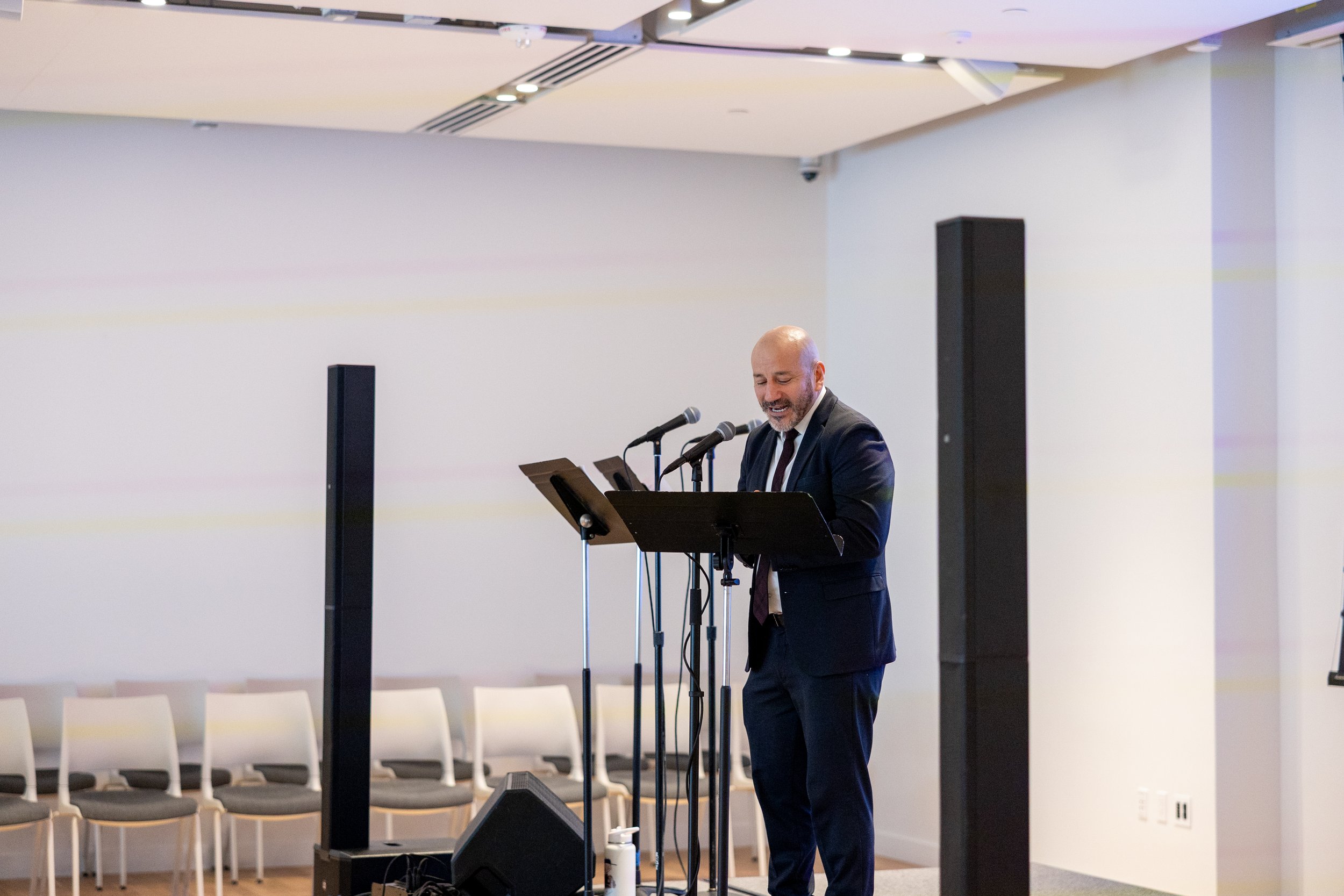 A man in a dark suit with a tie, standing at a podium with two microphones, speaks or reads from a paper in a conference room with rows of empty chairs.