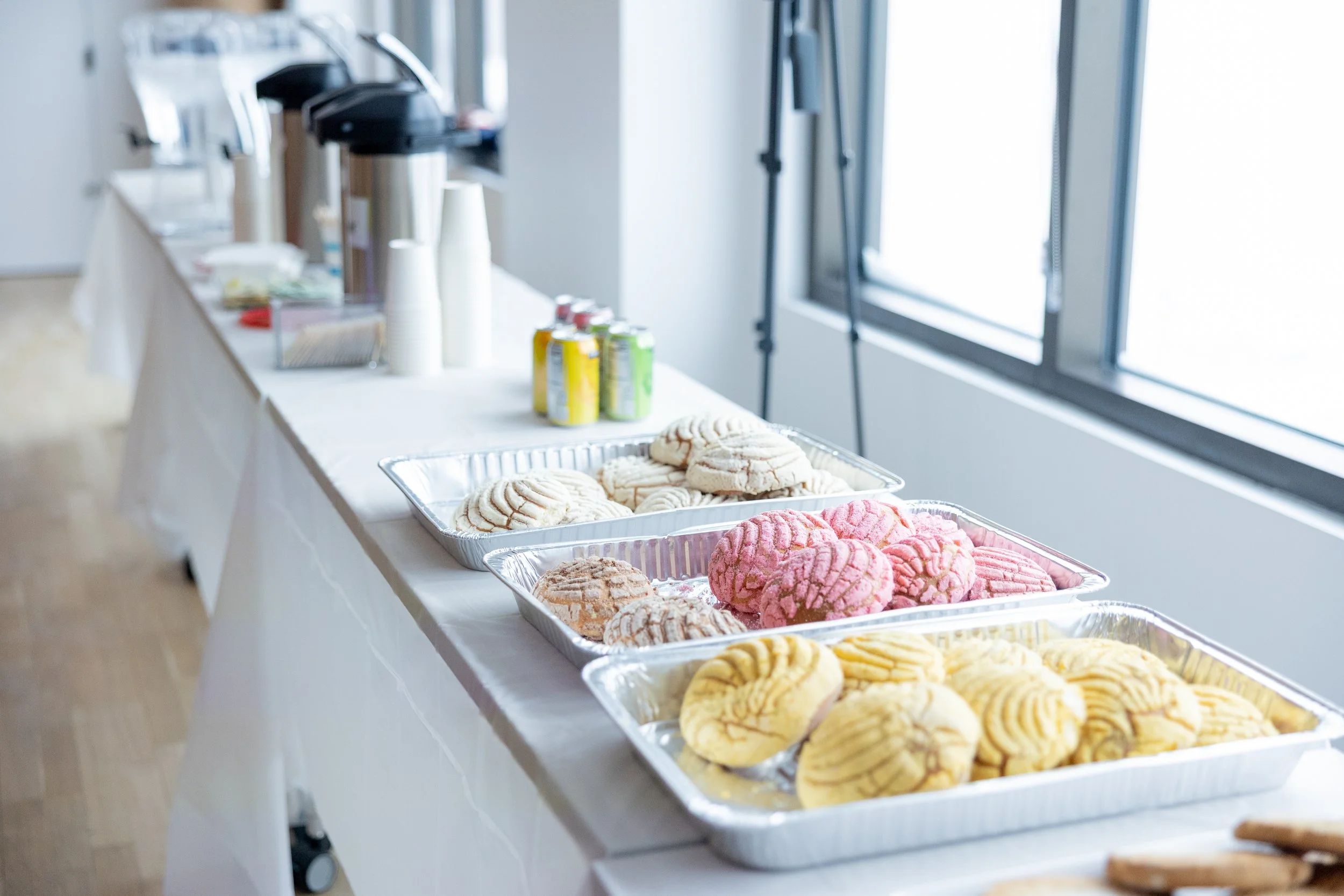 Assorted cookies on a long table near a window with coffee and soda cans in the background.
