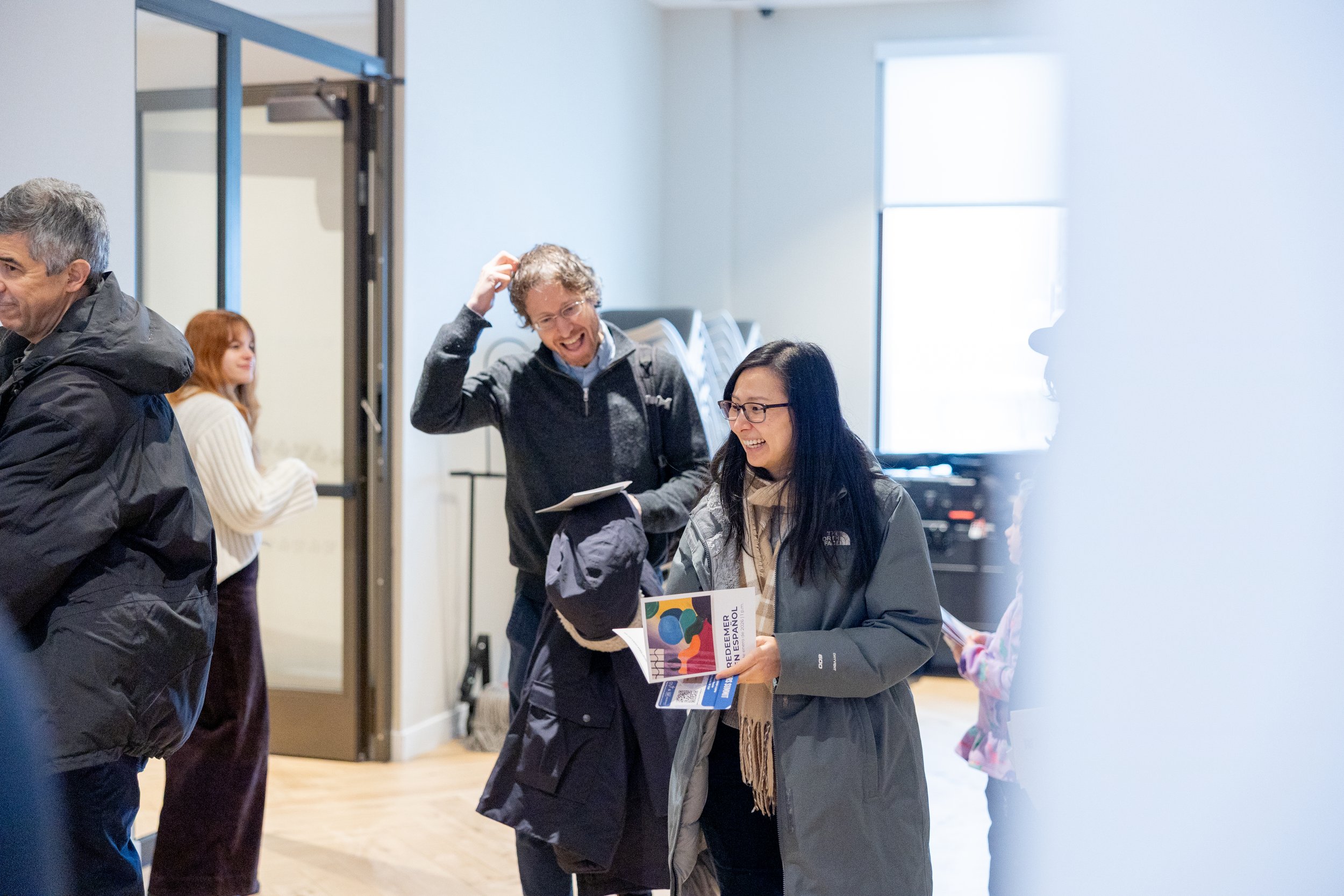 People inside a room, smiling and talking to each other. One woman holding a colorful brochure, a man with glasses laughing, another person partially visible, and a woman with red hair in the background near a door.