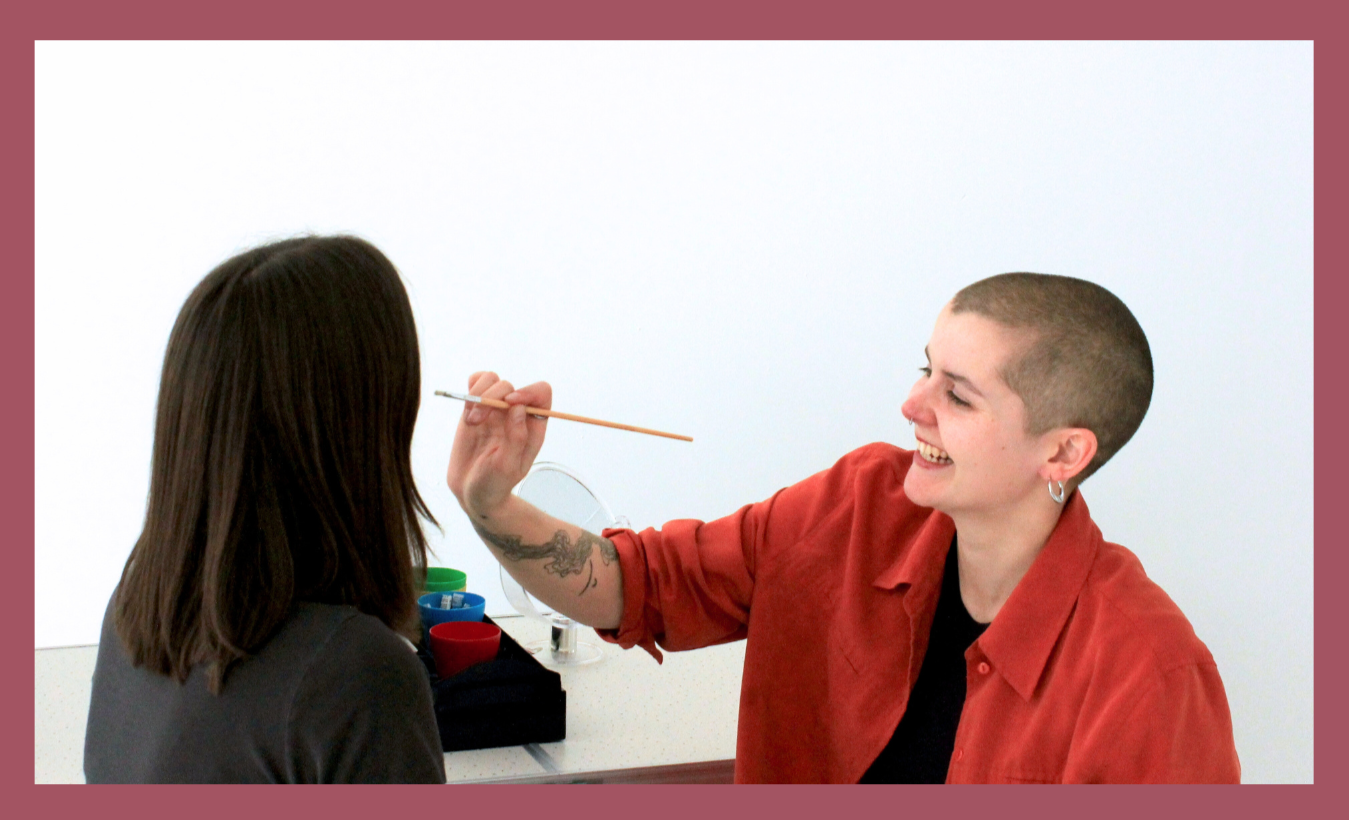 A smiling person holds a brush to a second persons face to face paint.