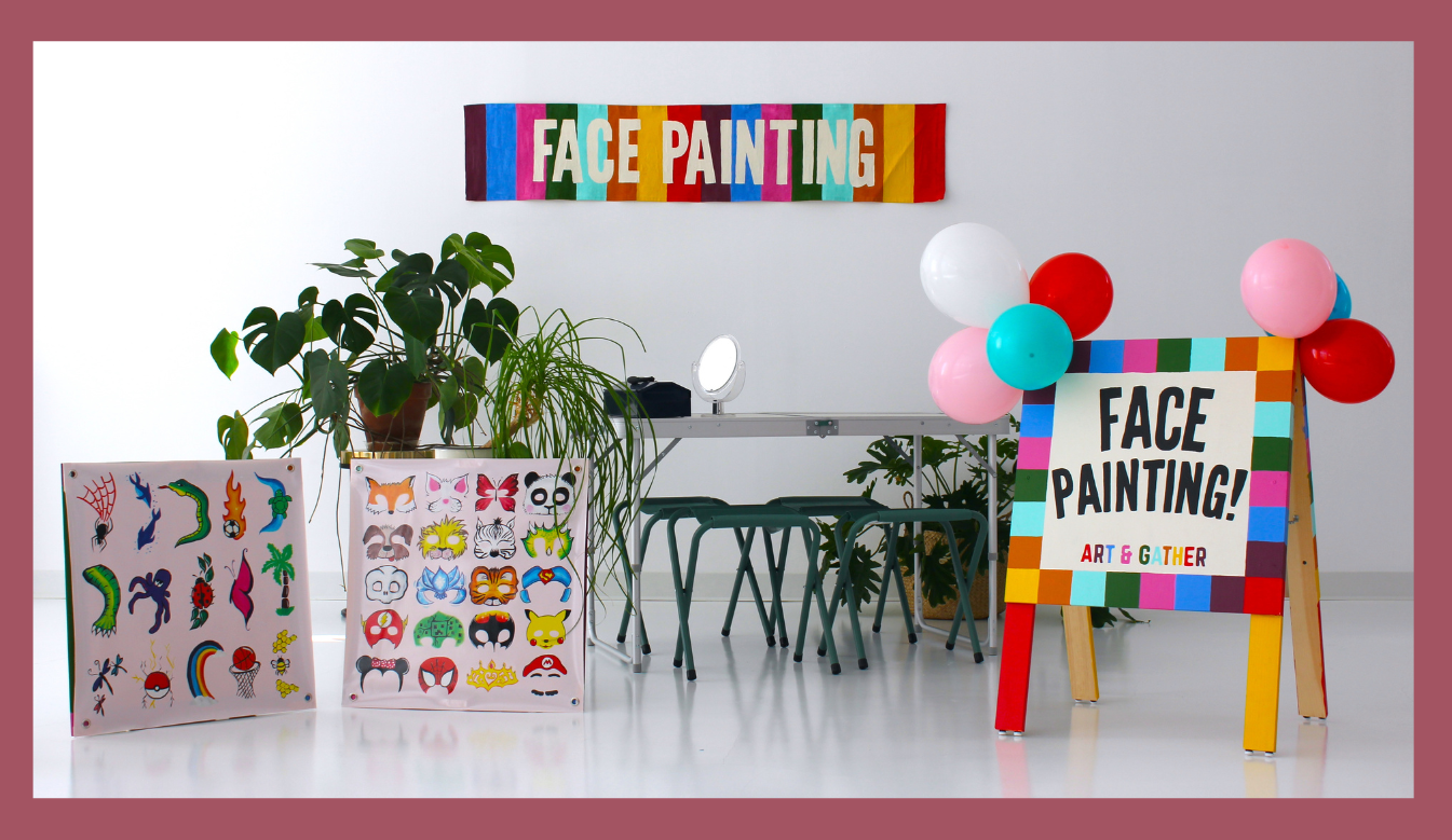 A white room with a colourful face painting booth set up. Plants are placed throughout the space. The set up includes a wall sign reading "face painting" two boards with facepainting examples, and a table and chairs.