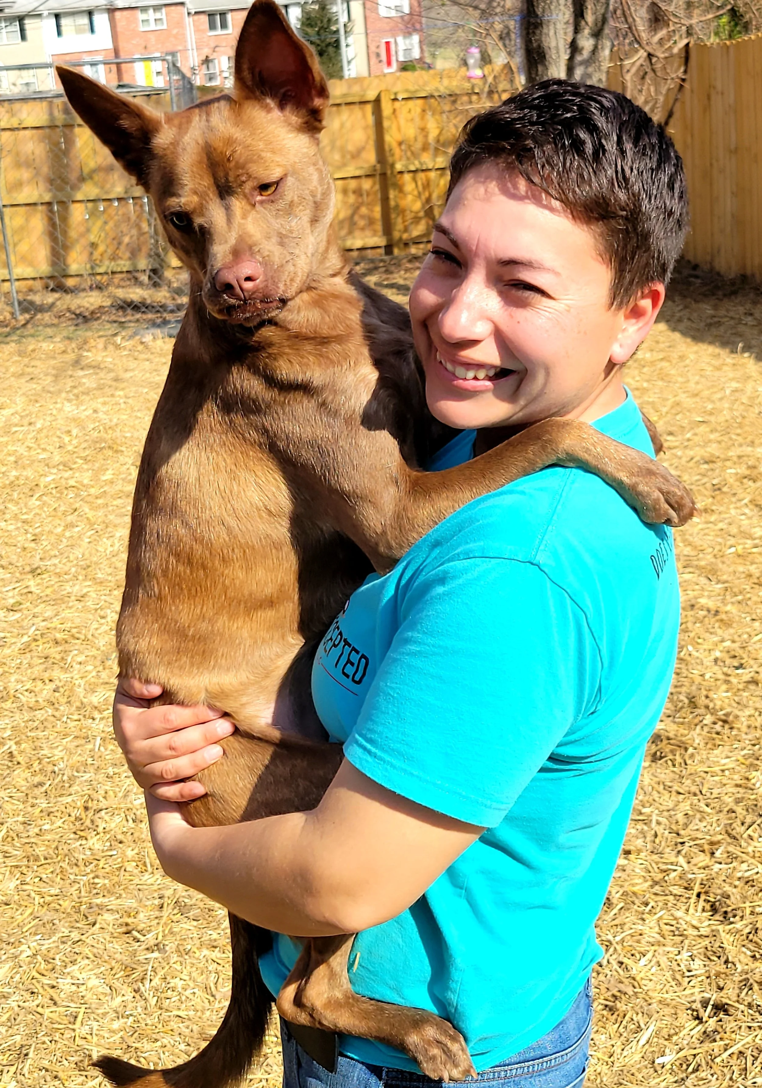 A person with short dark hair smiling and holding a brown dog with one eye eyes closed and one eye open, outside in a yard with a wooden fence, trees, and buildings in the background.
