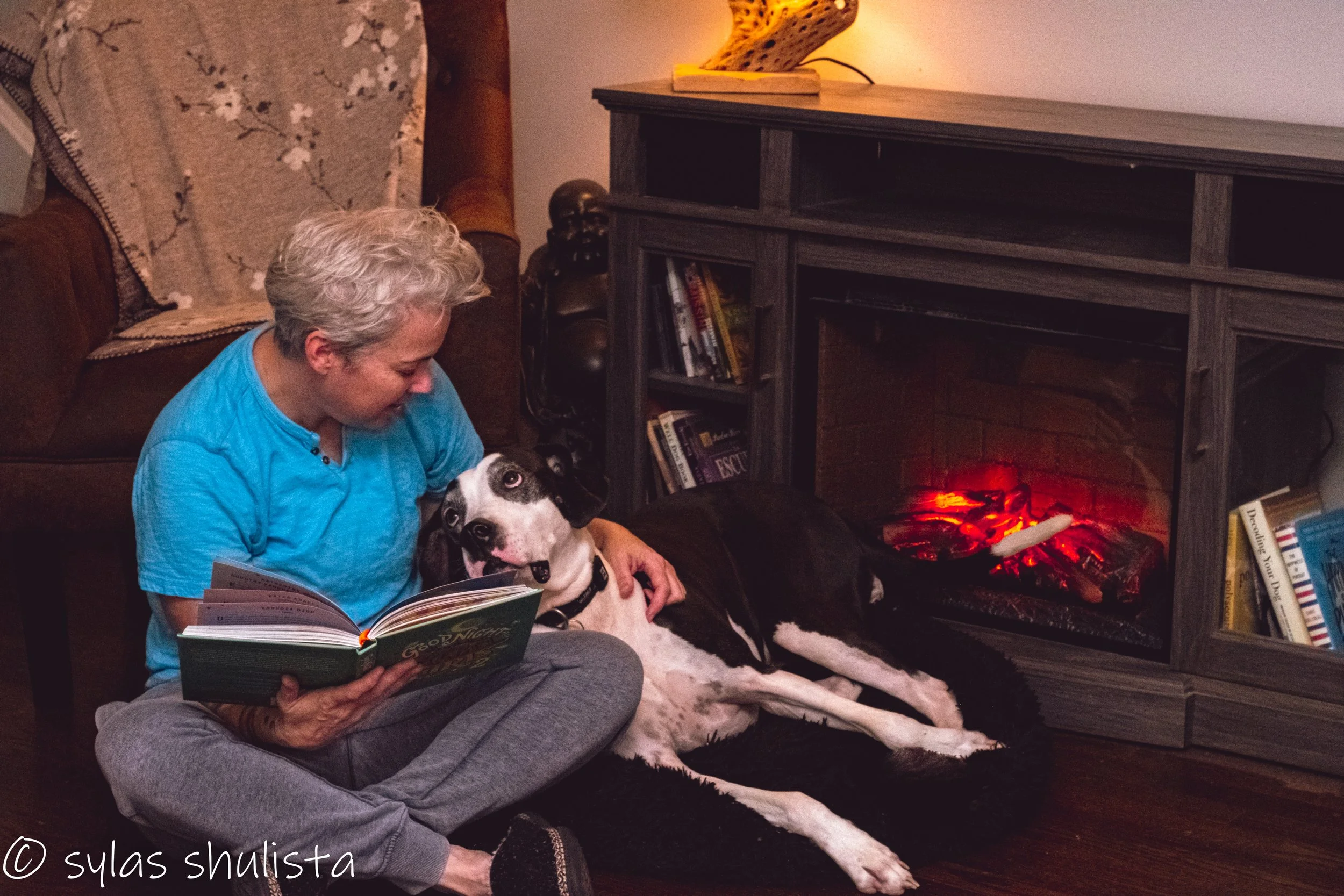 A woman with gray hair sitting on the floor reading a book and holding a large black and white dog, possibly a Great Dane, in front of a lit electric fireplace. There is a brown sofa with floral patterned pillows and a bookcase nearby.