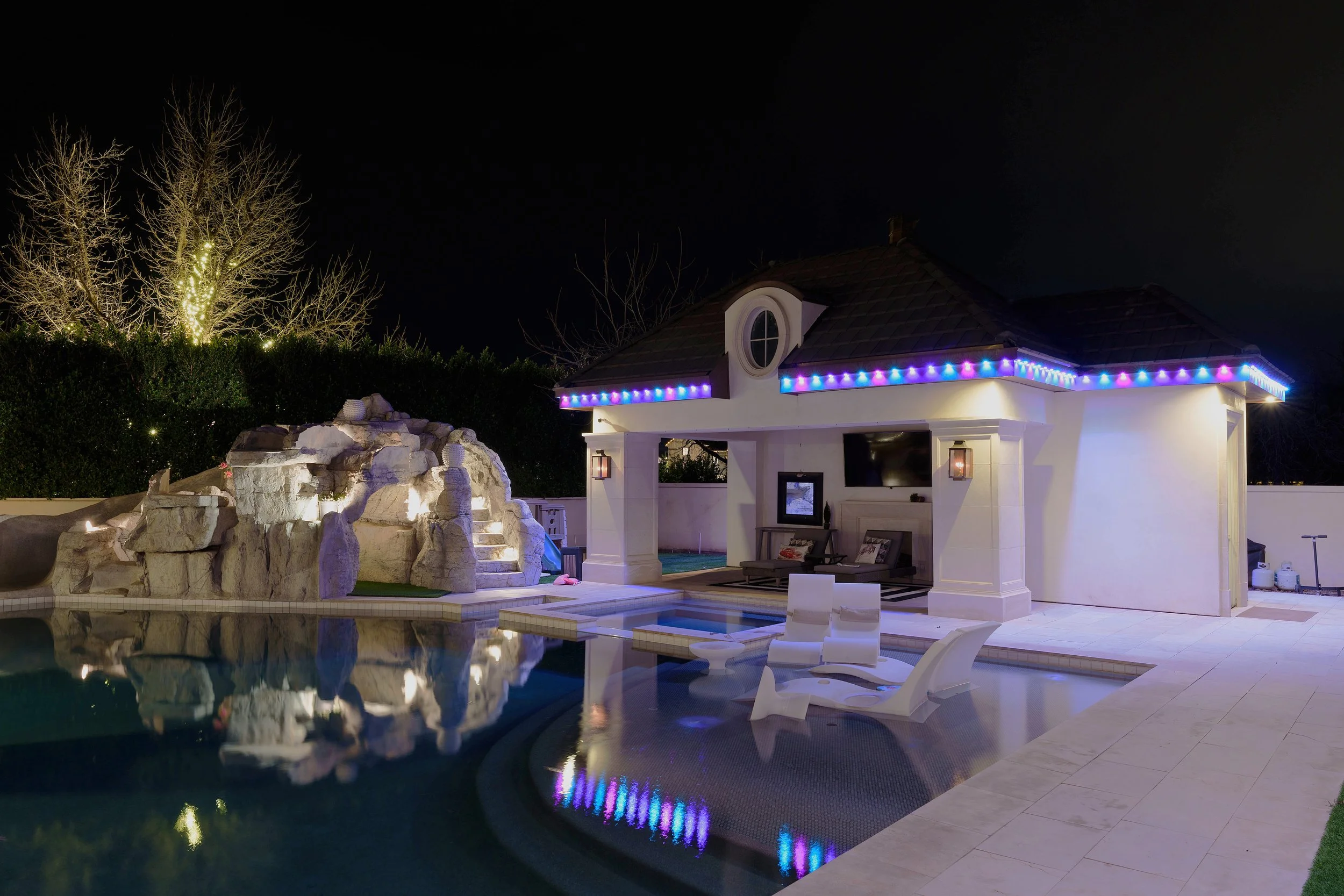 Nighttime view of a backyard with a swimming pool, stone waterfall feature, and patio furniture. A pool cabana with permanent LED lights along the roof edge and surrounding trees with lights are visible in the background.