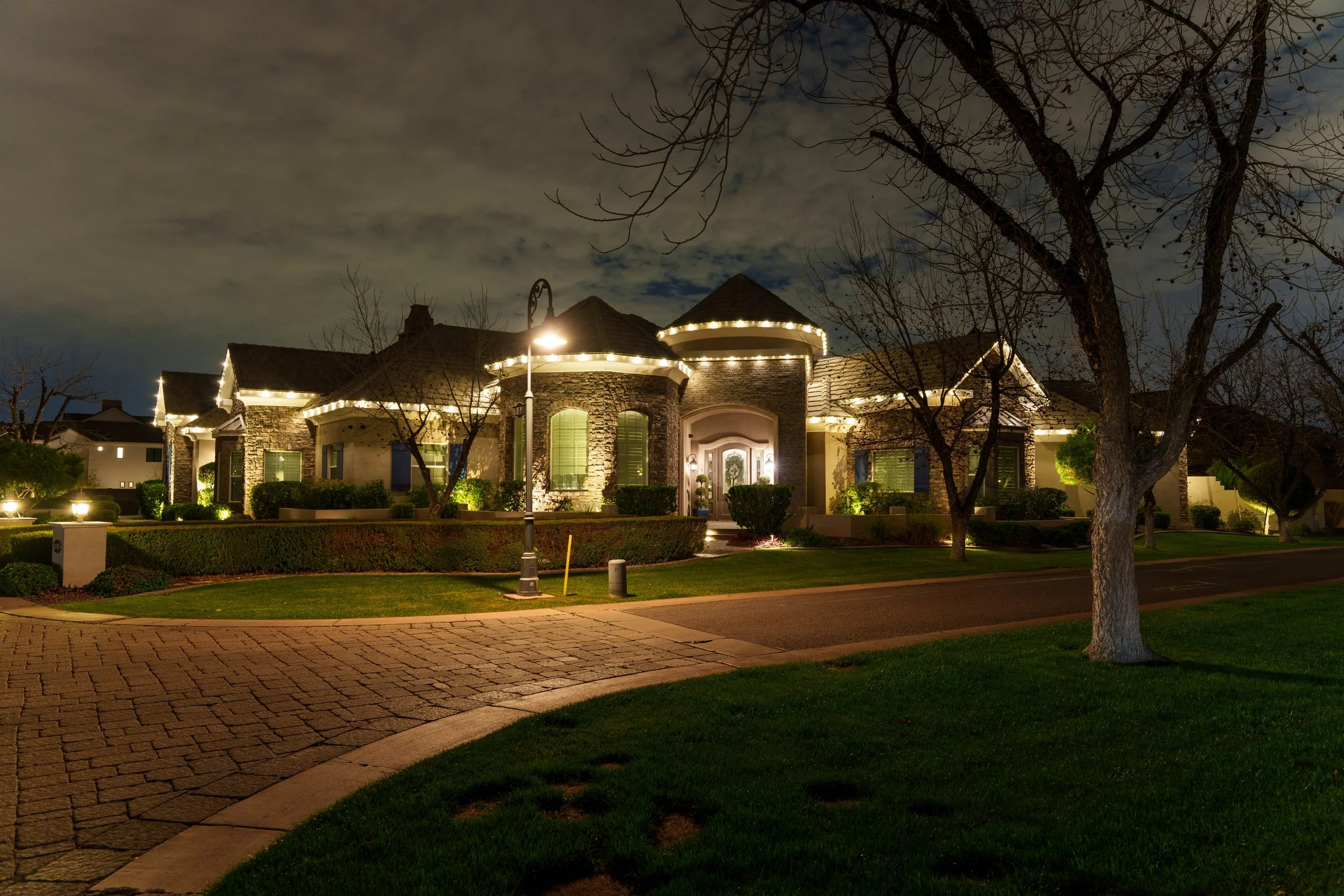 Nighttime view of a large house with stone walls and a steep shingled roof, illuminated with exterior LED lights. The front yard has well-manicured bushes, a leafless tree, and a paved sidewalk and street in the foreground. The sky is partly cloudy.