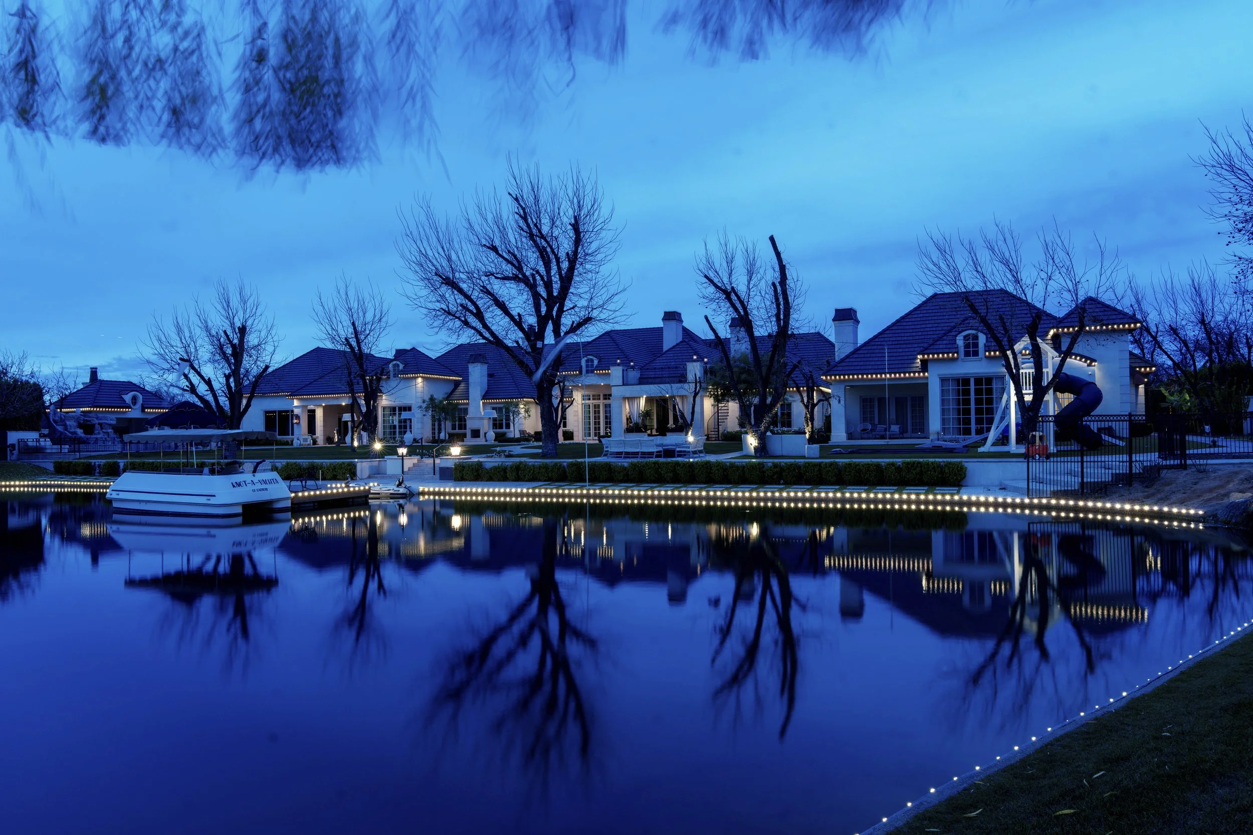 A high-end luxury home and casita lit with permanent roofline lighting, next to a calm body of water wrapped in permanent lights. A boat docked on the water, and leafless pecan atrees are in the background during dusk, under a partly cloudy sky.