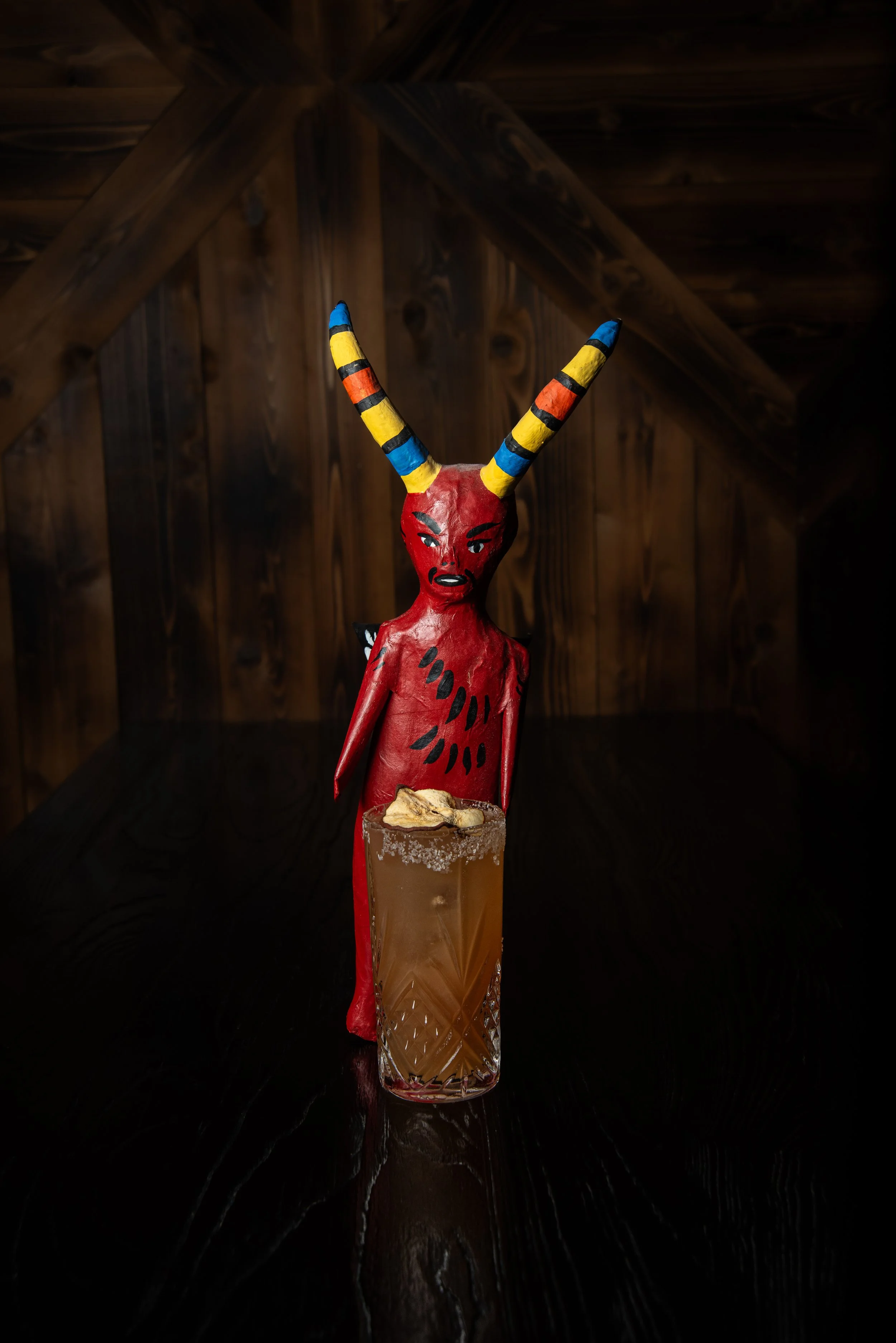 A glass of cocktail with salt on the rim, garnished with a thin slice of fruit, and a red figurine of a demon or devil with colorful striped horns, standing behind the glass.