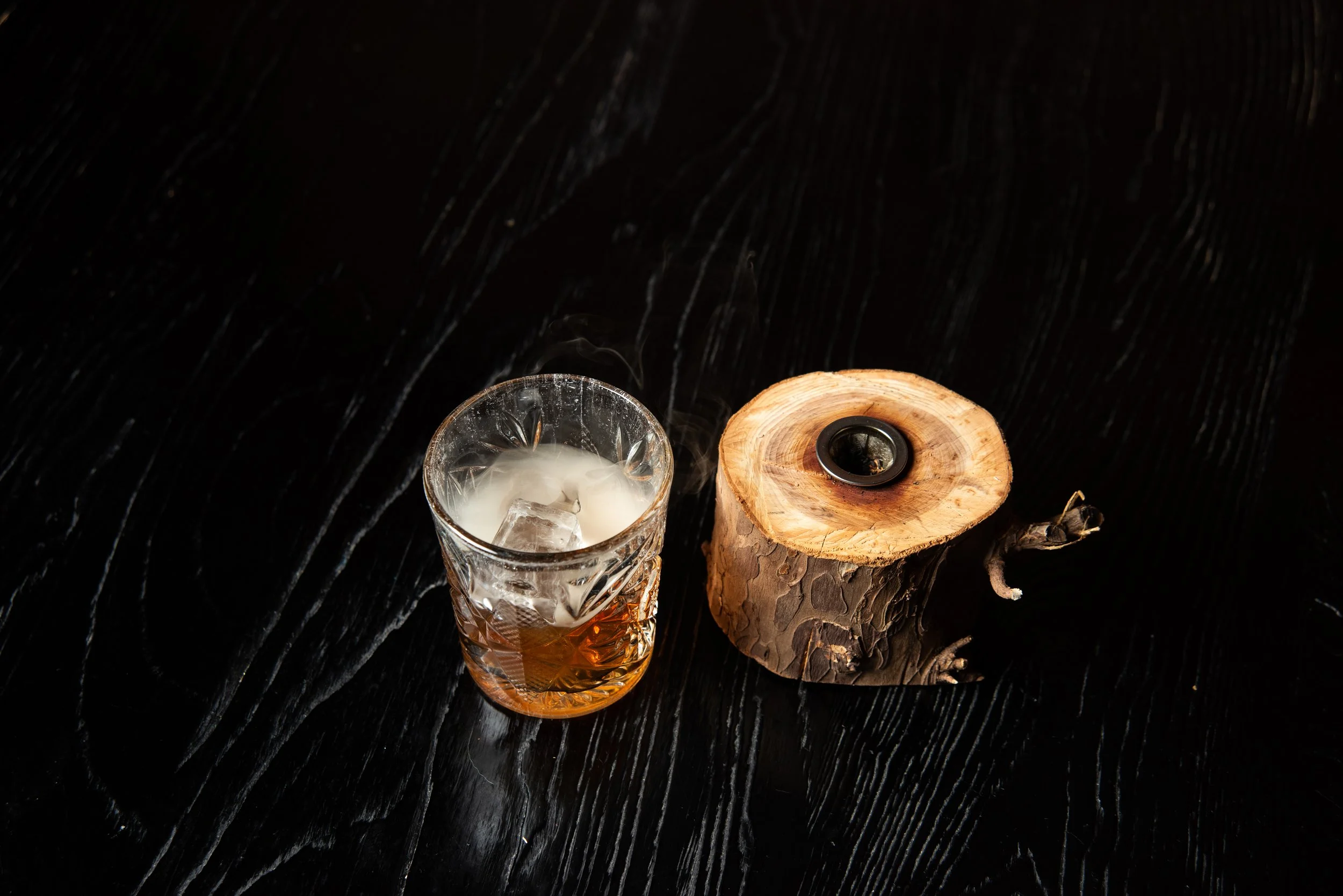 A glass of smoking whiskey cocktail with ice cubes and a burning candle on a black wooden surface.