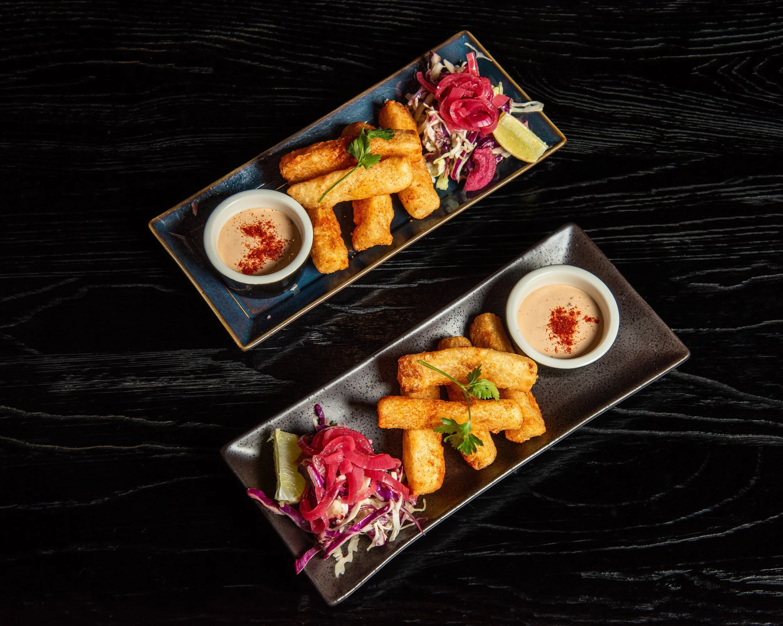 Two rectangular plates with fried yucca fries, cabbage, and dipping sauce on a dark wooden table.