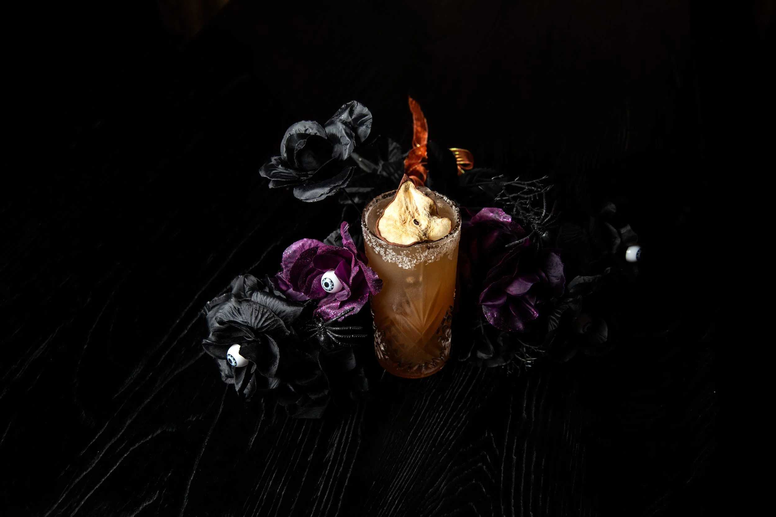 A Halloween-themed drink in a detailed glass, garnished with a dried pear slice and surrounded by black and purple artificial flowers with eye decorations, set on a dark wooden surface.