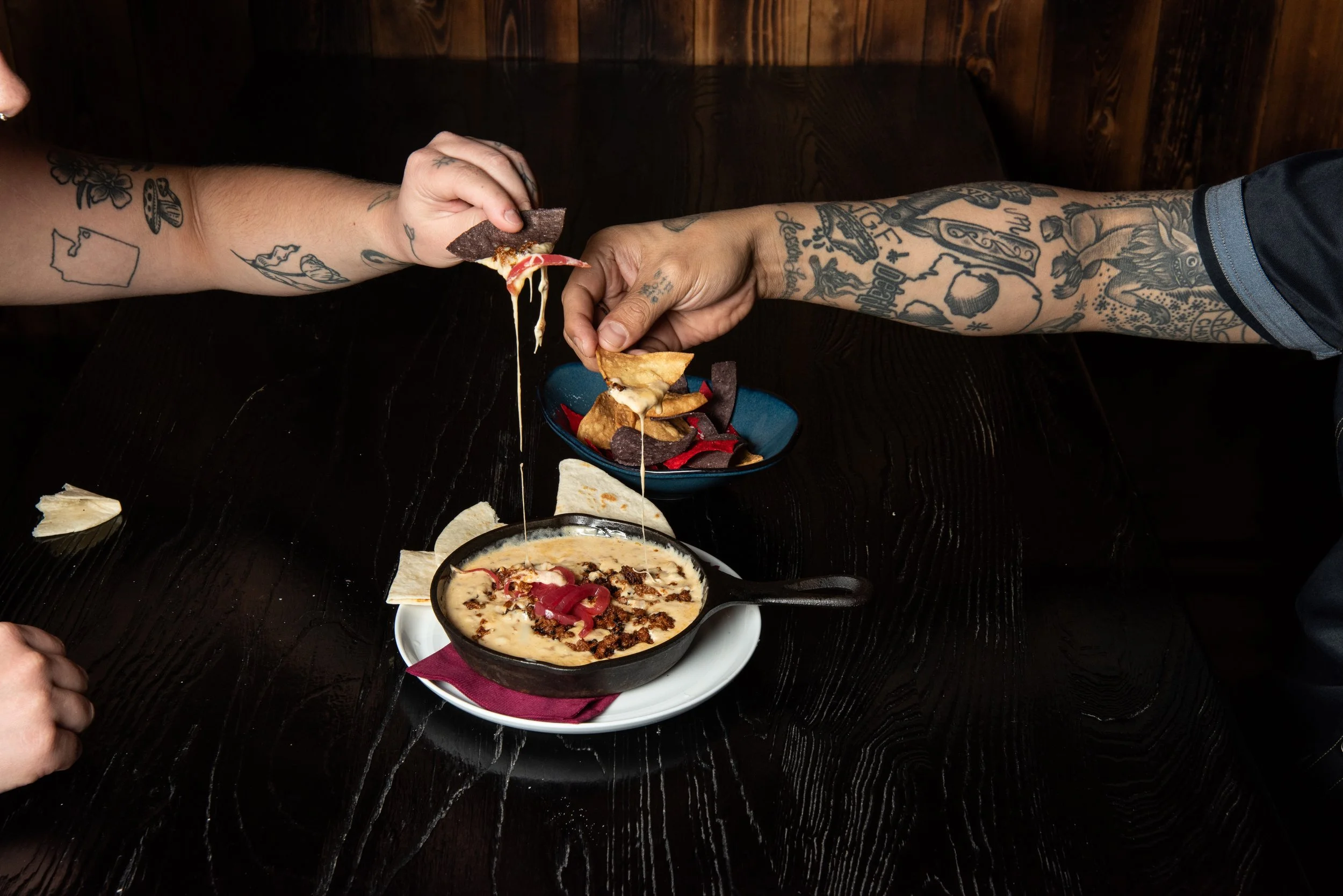 Two tattooed hands sharing a cheese dip with chorizo and  chips at a dark wooden table at a Capitol Hill mexican restaurant
