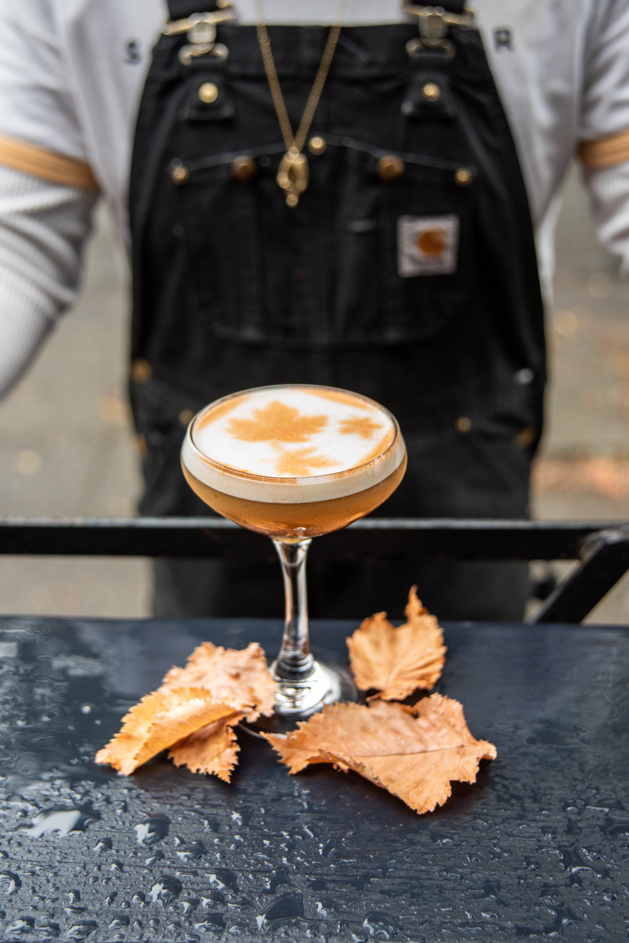 A cocktail with foam and leaf-shaped decoration on top, placed on a black wet surface with fallen autumn leaves around the glass, in front of a person wearing a black Carhartt overalls and a gray shirt.