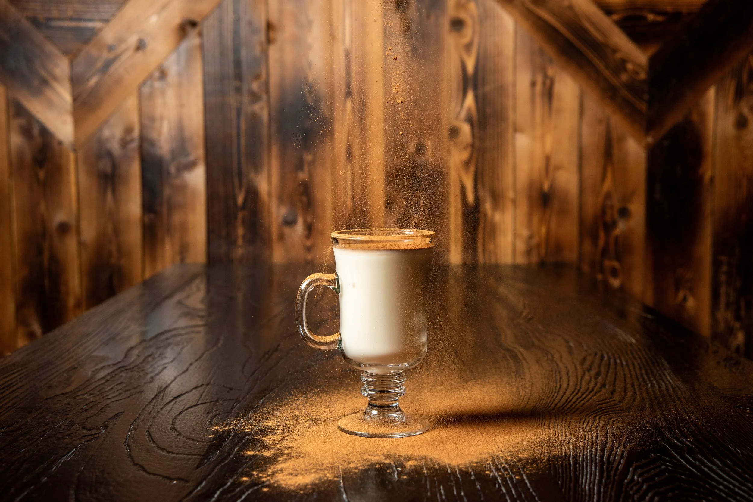 A glass mug filled with atole, a corn/masa based warm drink, on a dark wooden table, with a powder or dust falling into it, creating a cloud of dust around the mug, against a wooden paneled background. Aesthetic Seattle Capitol Hill Cocktails