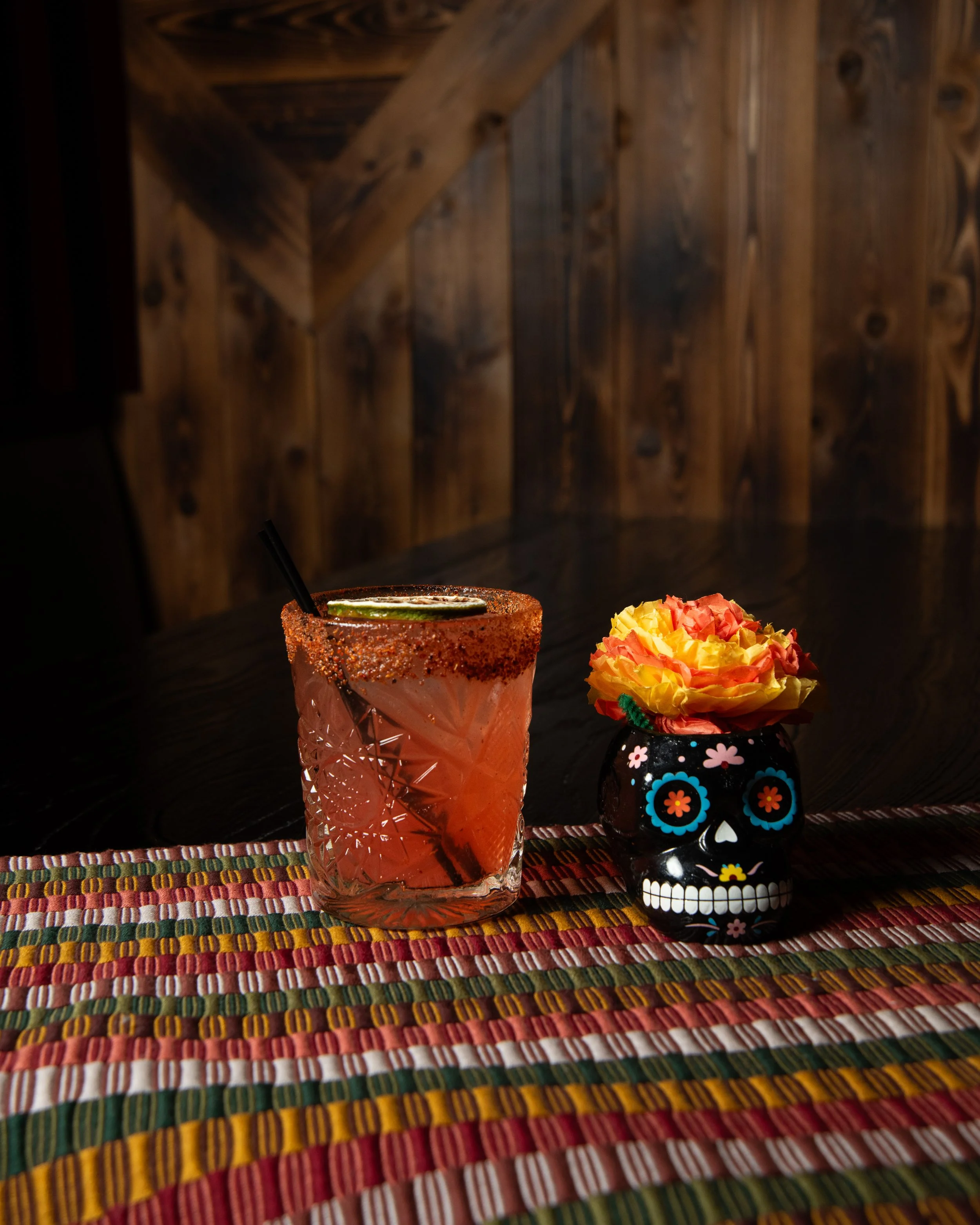 A colorful Mexican style table setting with a pink cocktail garnished with lemon and lime slices, and a small black skull-shaped vase with a yellow and pink paper flower, all placed on a woven multicolored table runner with a wooden wall background.