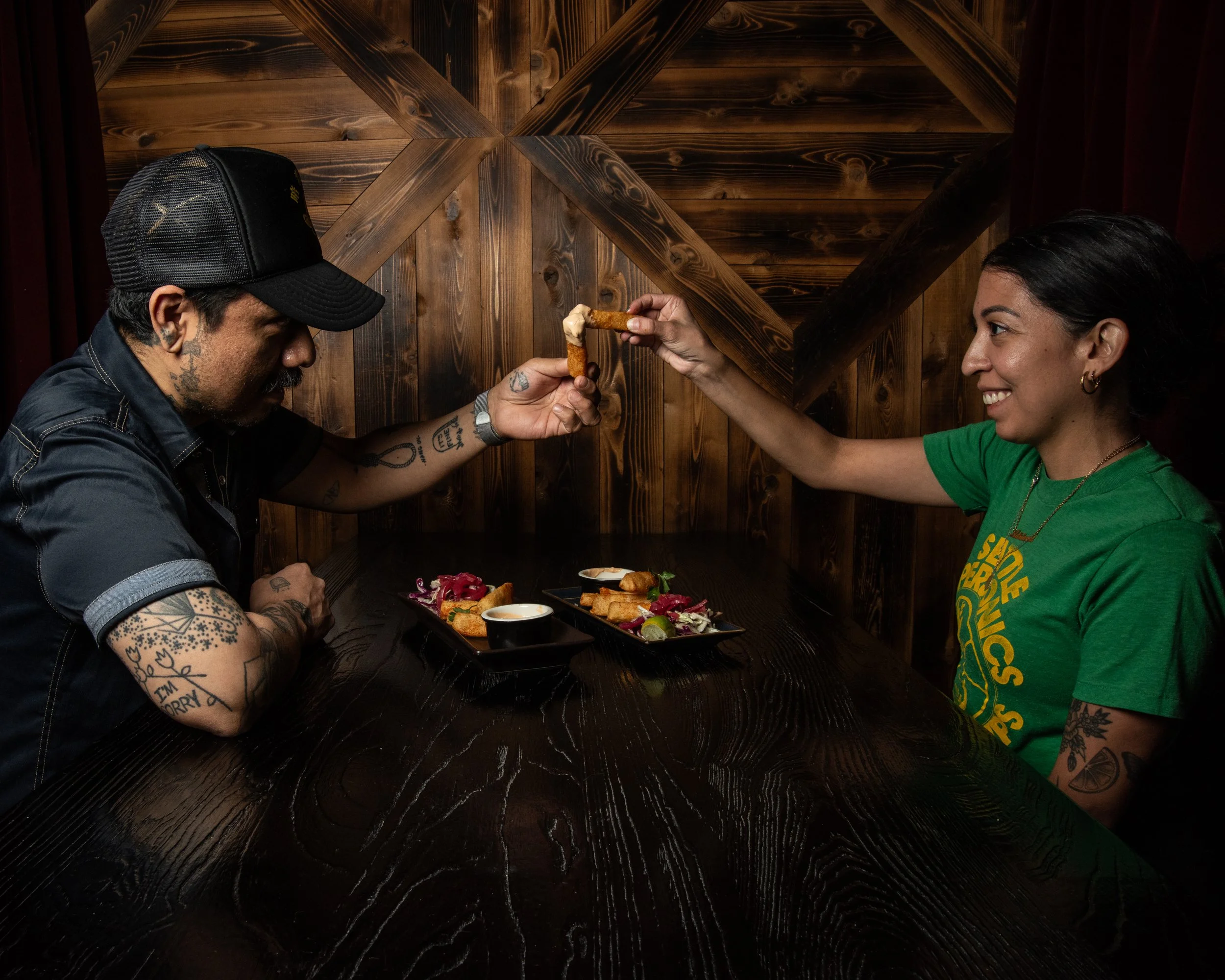 A man and woman are sitting at a dark wooden table in a restaurant with wooden walls. The woman is smiling and offering queso fundido, authentic mexican cheese. Mystery Seattle Capitol Hill cocktail goth Bar with Mexican Tapas & Food, vegan options