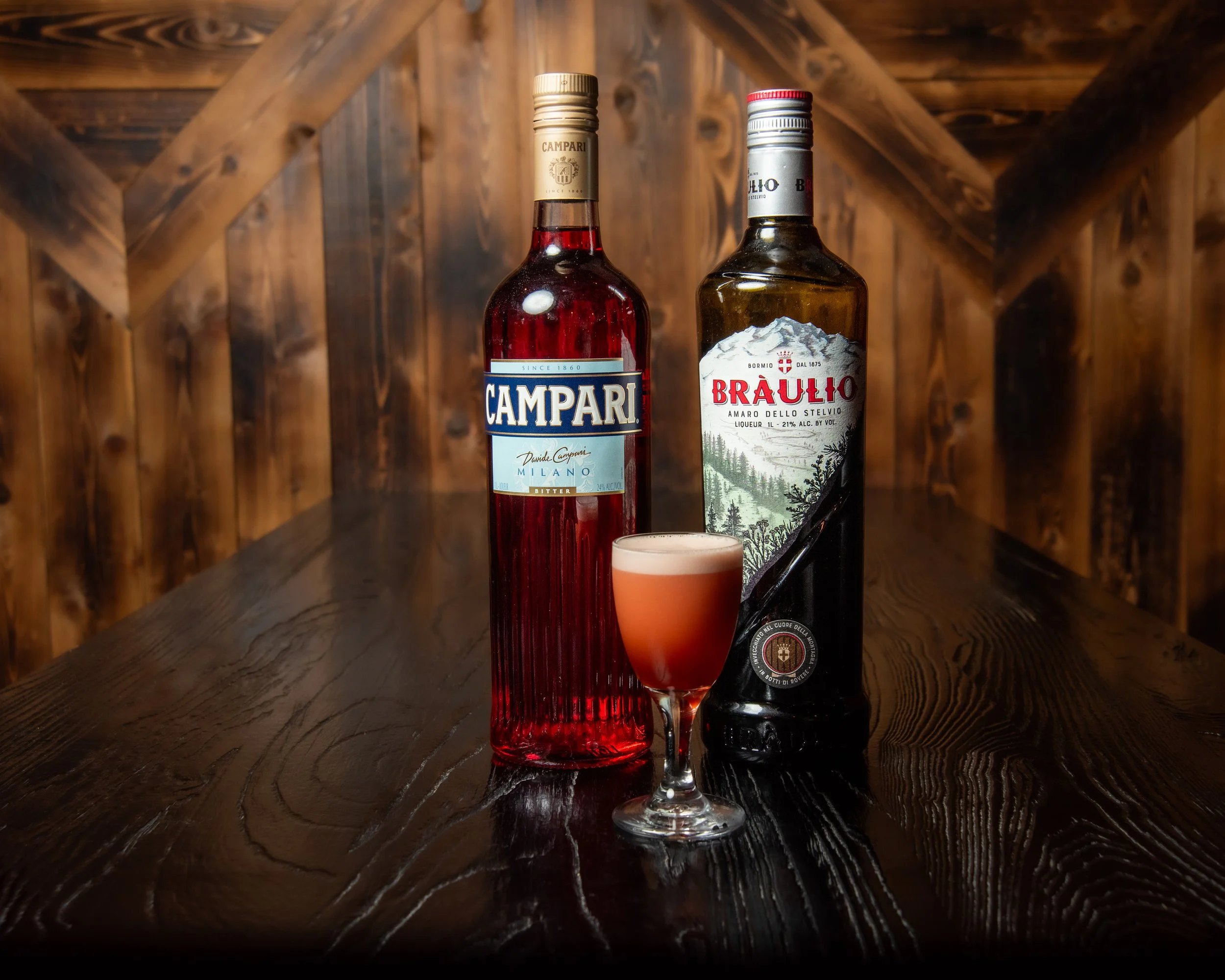 Two bottles of Italian liquor, Campari and Bàulio, along with a cocktail glass filled with a pink-colored drink on a dark wooden table in a rustic wooden interior. Craft cocktails in a vibey capitol hill bar.
