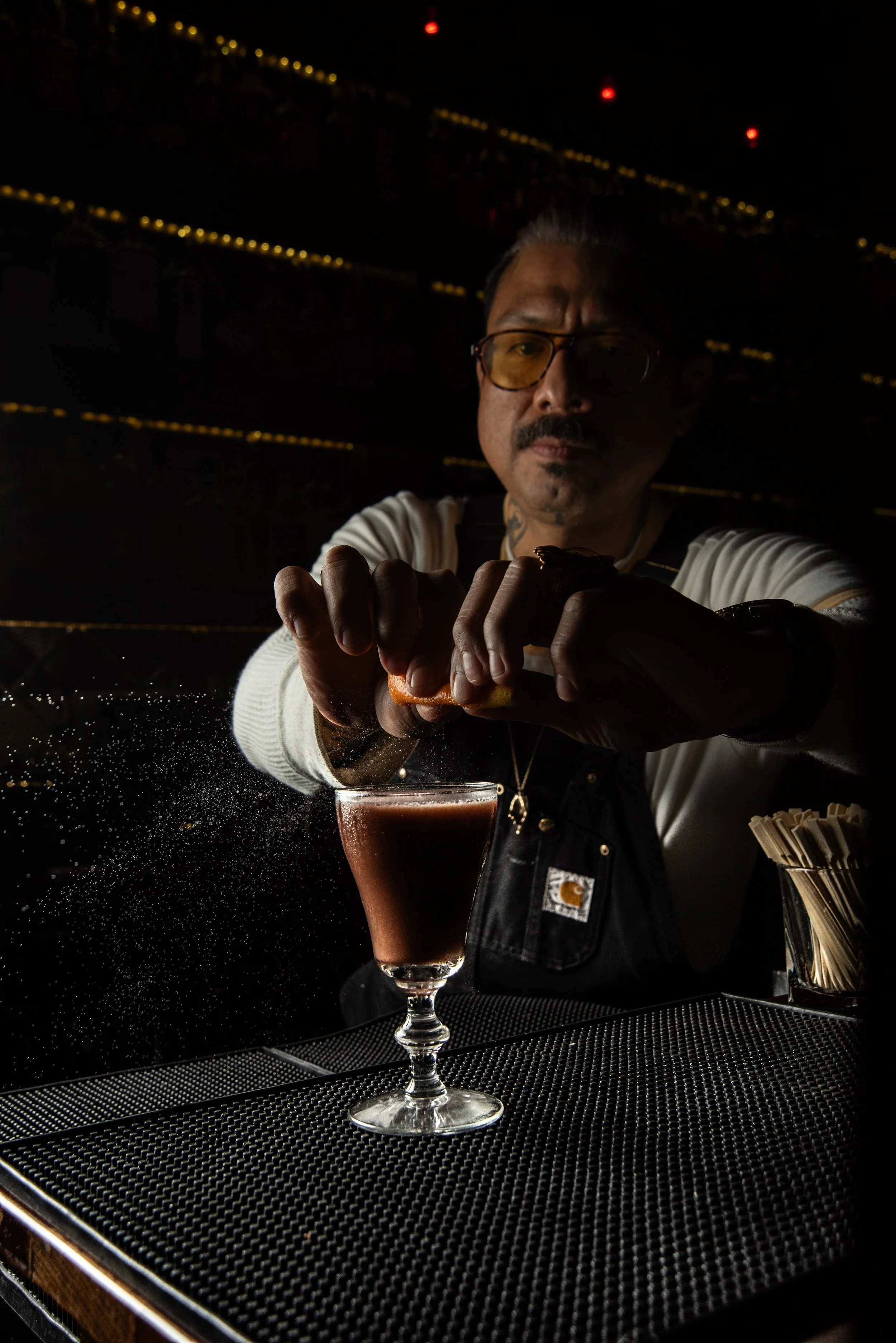 Bartender squeezing a citrus fruit into a brown cocktail in a glass, with a dark background and bar setting.