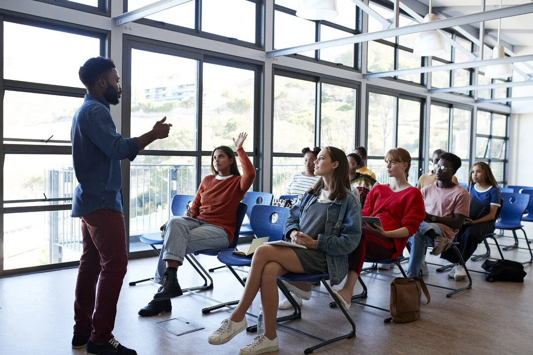 A diverse group of young adults sitting in a large, bright room with floor-to-ceiling windows, listening to a man speaking at the front of the room.