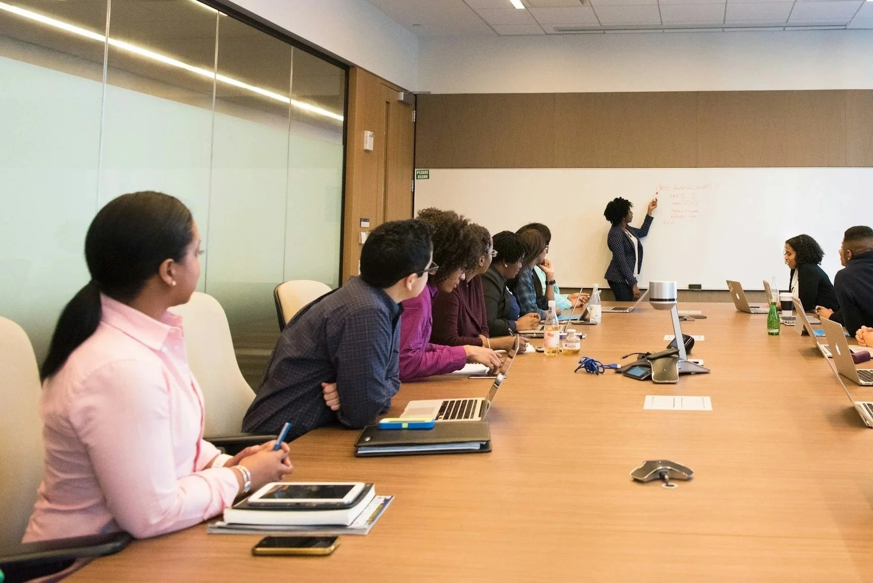 A diverse group of people is attending a meeting or presentation in a modern conference room. One woman is writing on a whiteboard at the front, while others are seated at a long table with laptops, notebooks, and drinks.